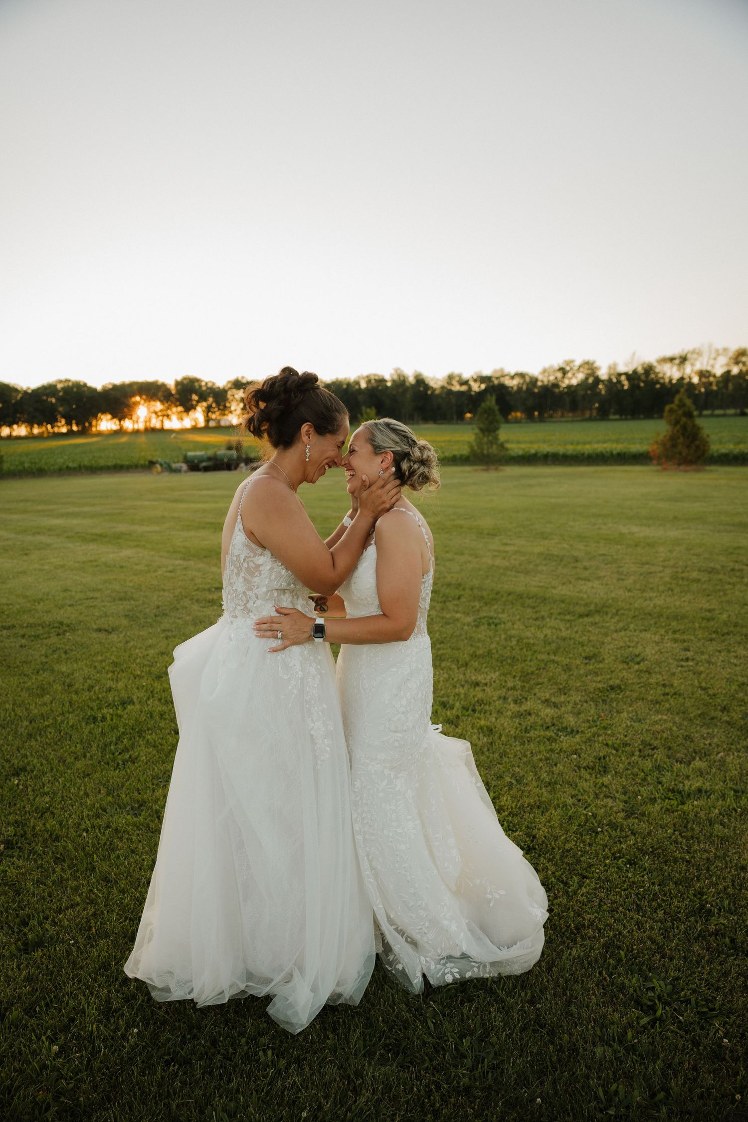 Two brides in white lace gowns share an intimate moment at sunset on a lush green lawn.