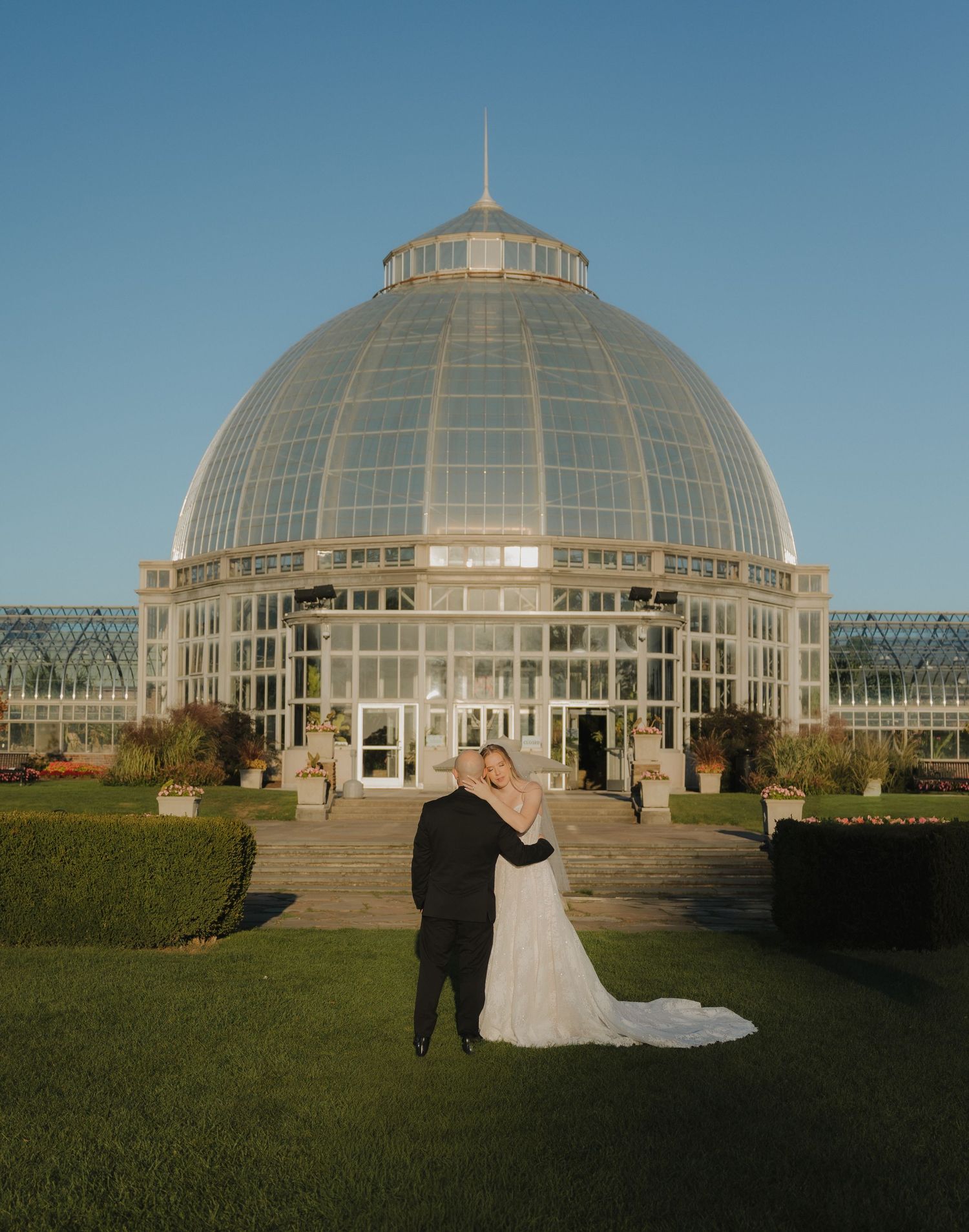Bride and groom embrace before a grand glass dome conservatory on a sunny day at Belle Isle.