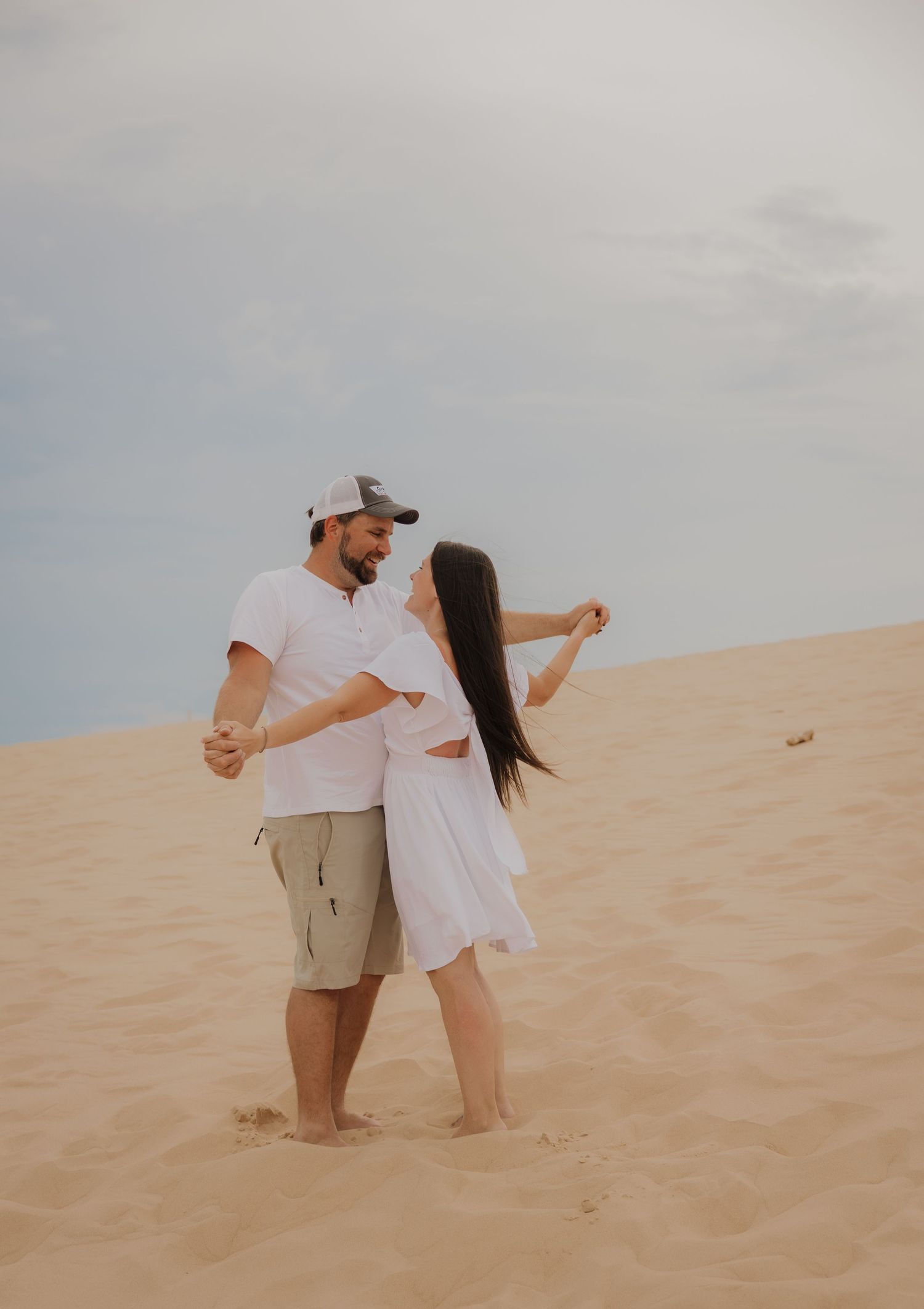 A couple dances barefoot on sandy dunes, woman in white dress, man in white shirt and cap, under cloudy sky.