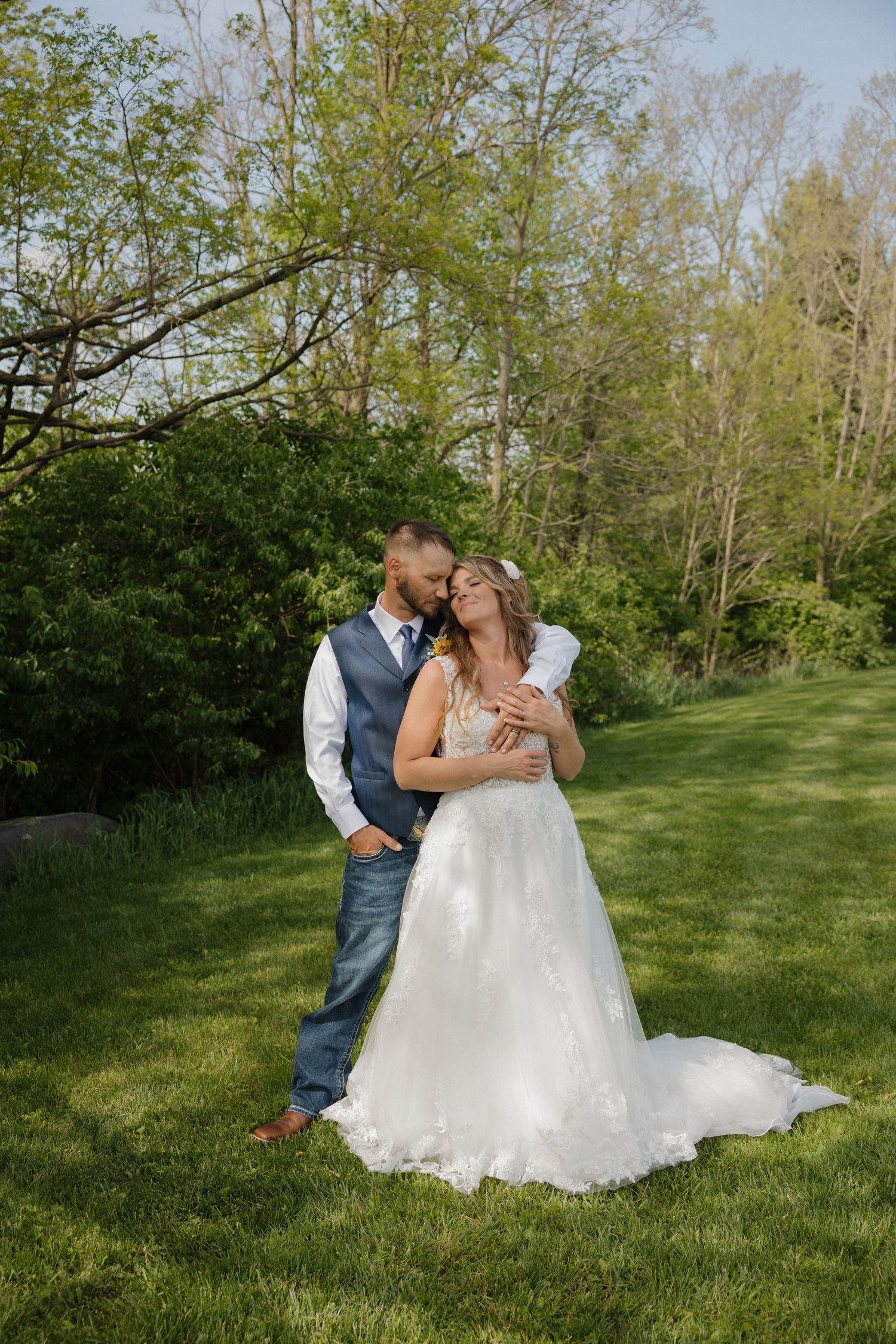 Bride and groom embracing outdoors on lush green lawn surrounded by trees on their wedding day.