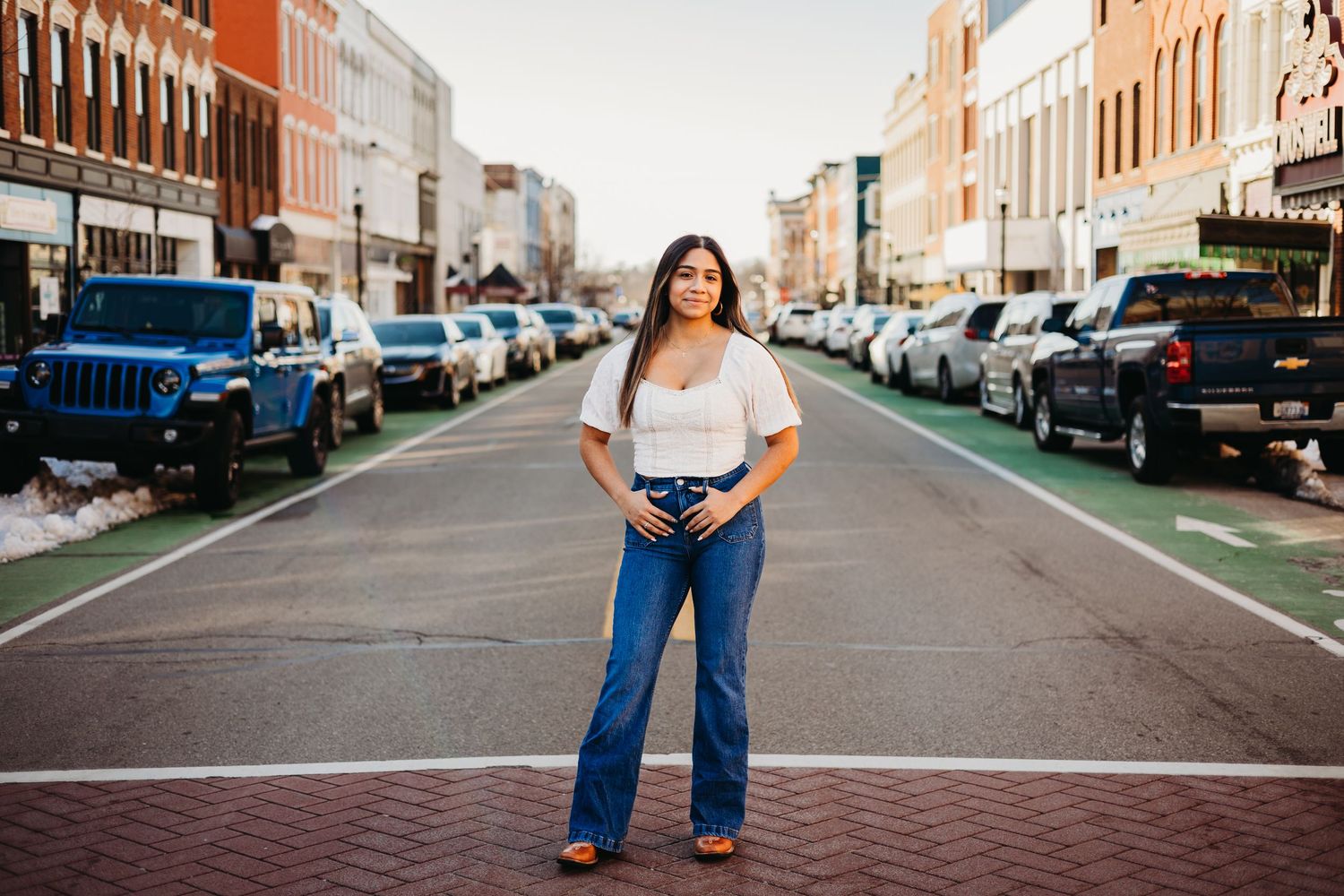 Young woman in white top and flared jeans poses confidently on a brick downtown street lined with parked cars.