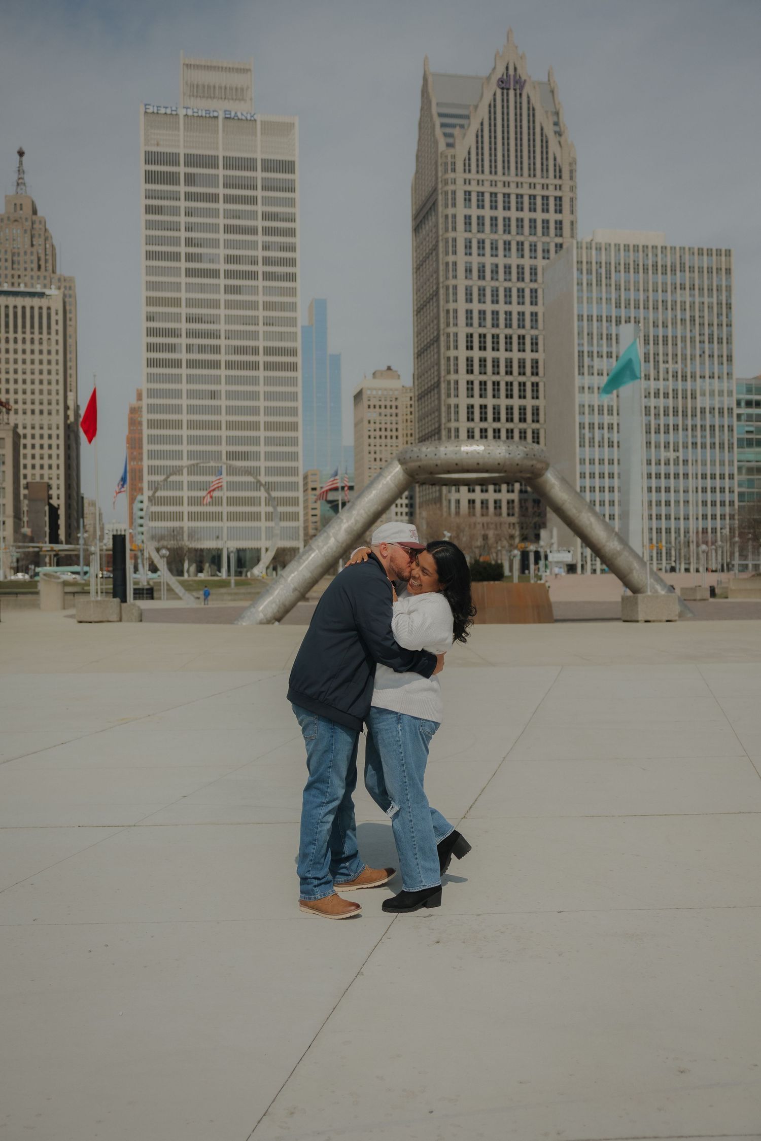 Couple sharing a romantic kiss at Hart Plaza in Detroit with iconic city skyline and the Transcending sculpture in the background.