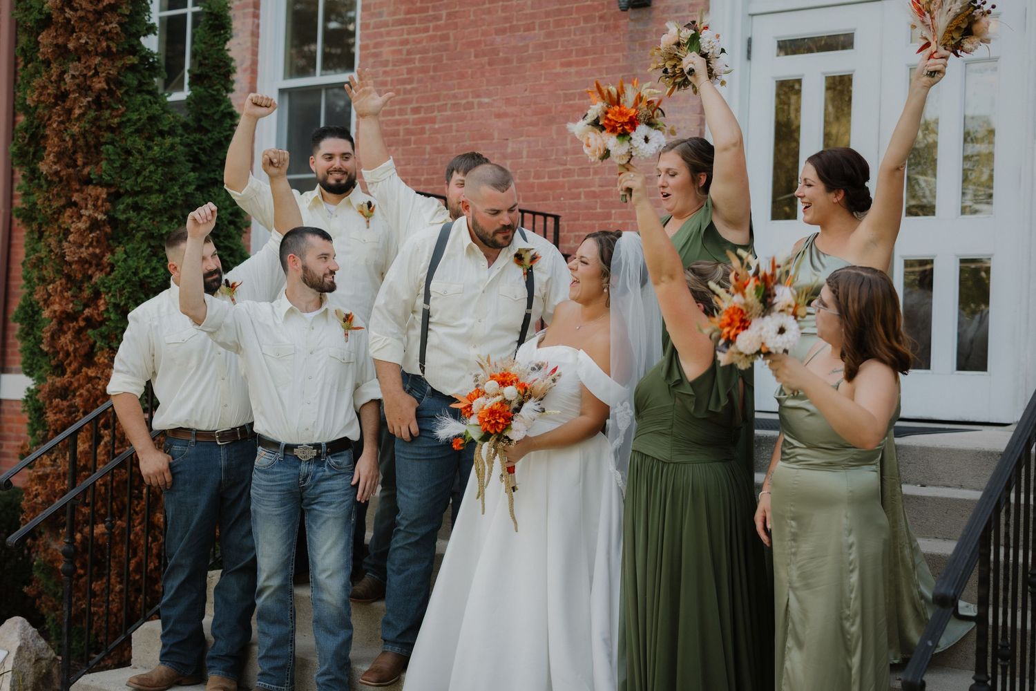 Wedding party celebrating on steps, bride in white gown with orange bouquet, groomsmen in jeans, bridesmaids in sage green dresses.