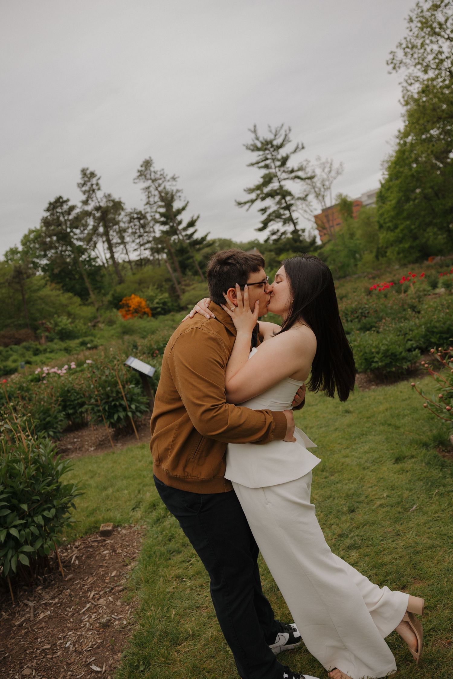 Couple sharing a romantic kiss while embracing in a lush garden setting on an overcast day.