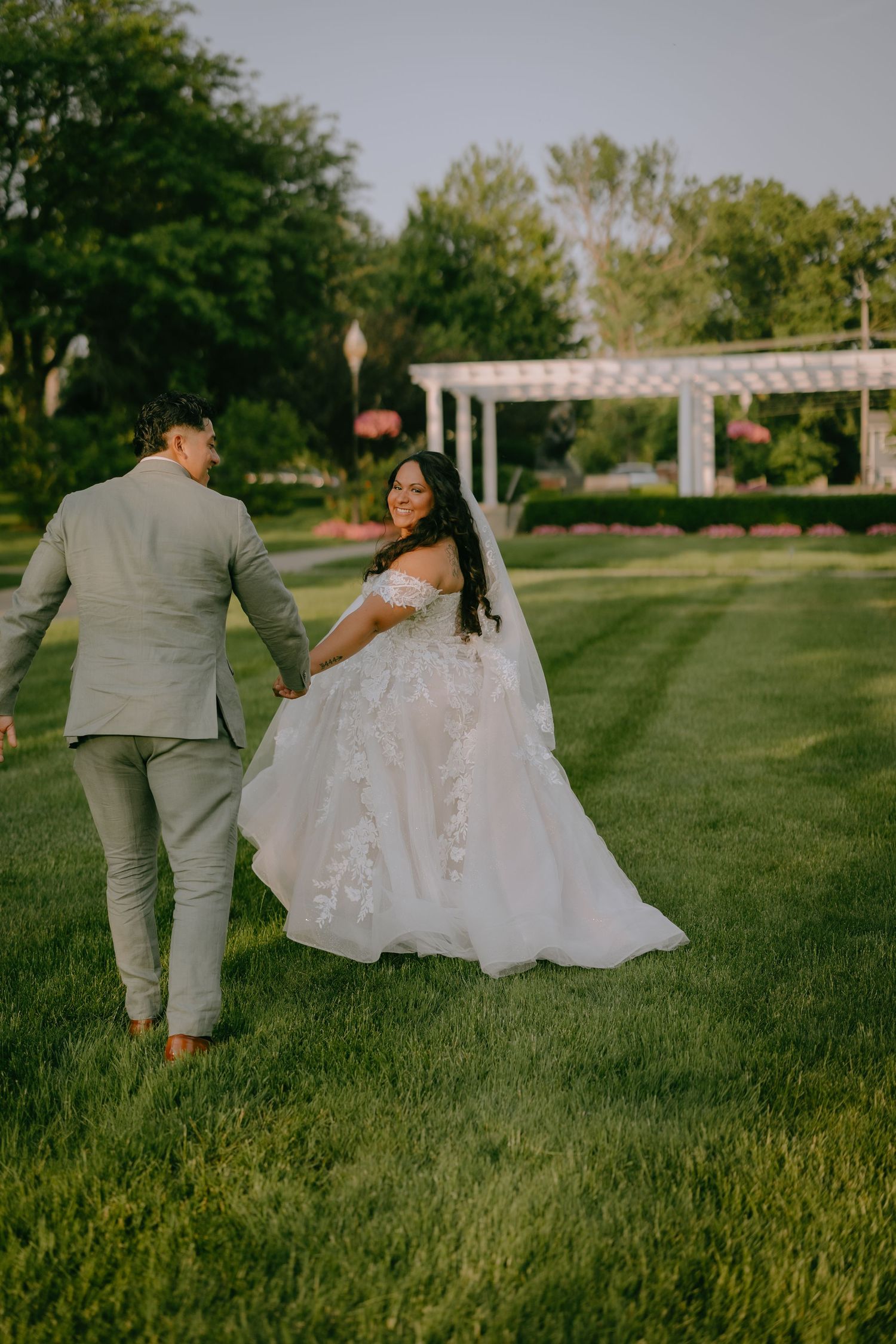 A bride in a flowing lace gown and groom in grey suit share a romantic moment on manicured garden lawn.