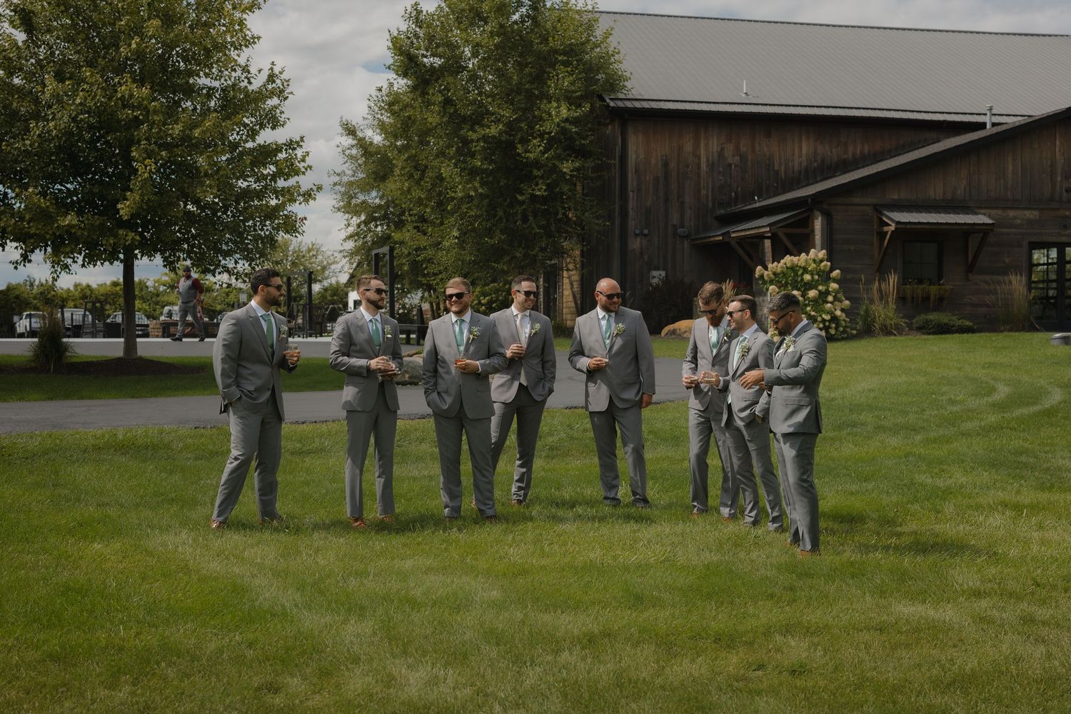 Wedding party in gray suits stands together on a grassy lawn during an outdoor celebration.