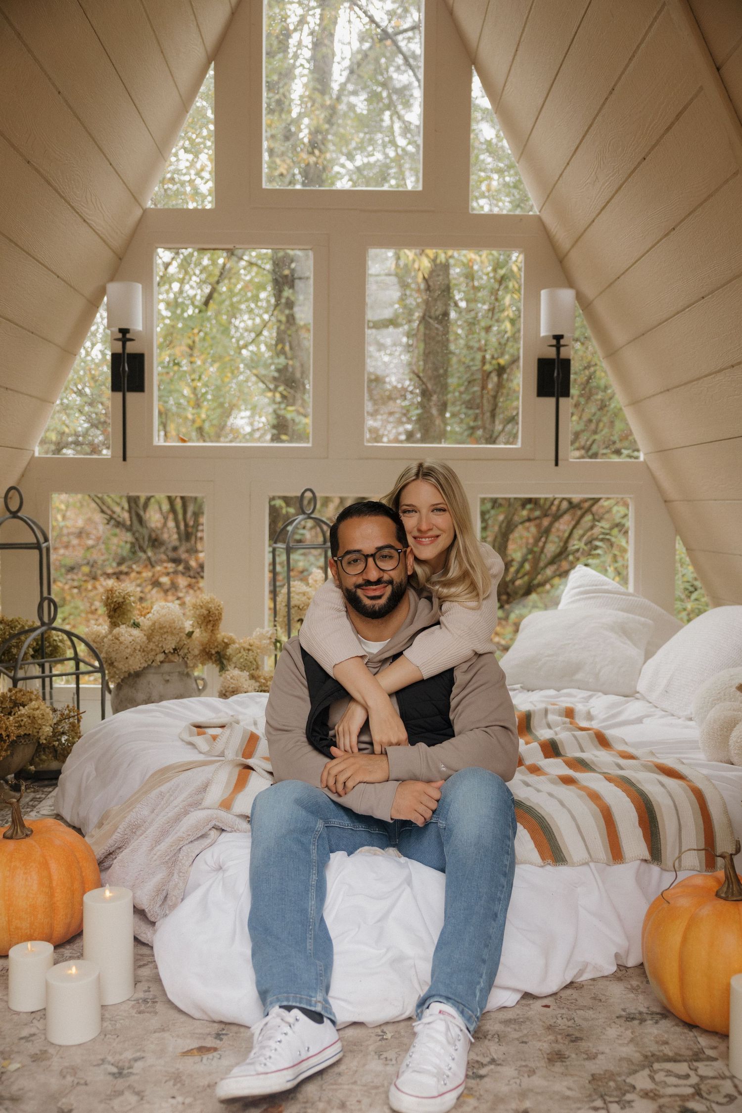 A cozy indoor scene with throw pillows, white blankets, and orange pumpkins arranged in a bright sunlit room.