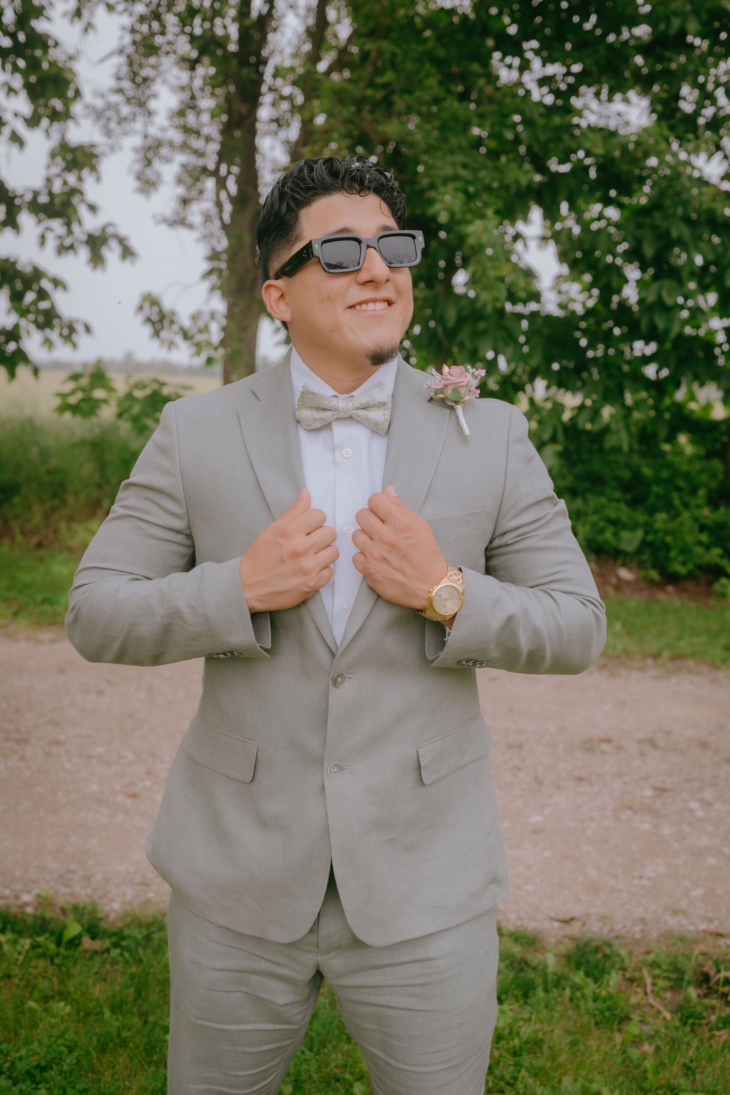 A person in a light gray suit and bowtie smiles while adjusting their jacket lapels outdoors on a sunny day.