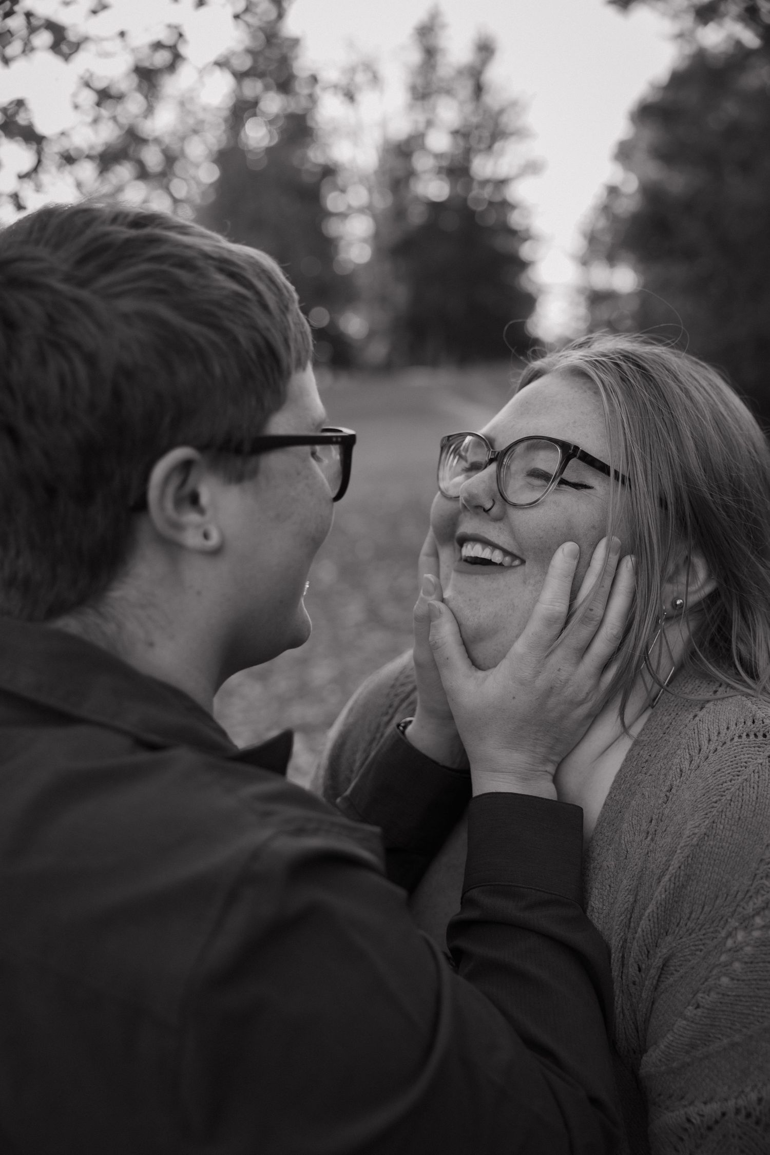 Black and white photo of a couple laughing, man tenderly holding woman's face, both wearing glasses outdoors.