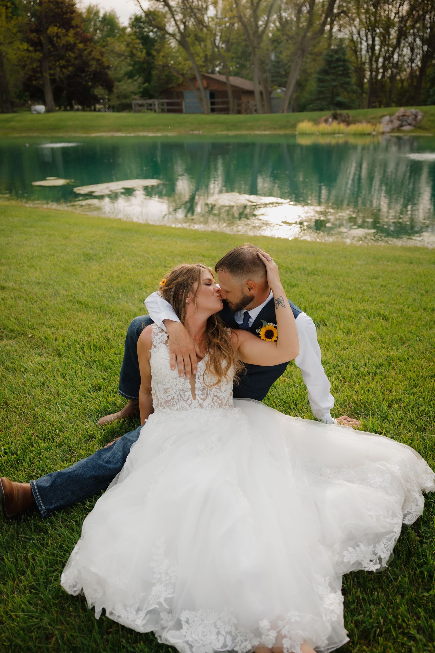 Bride and groom share a romantic kiss sitting on green grass beside a serene pond at their outdoor wedding.