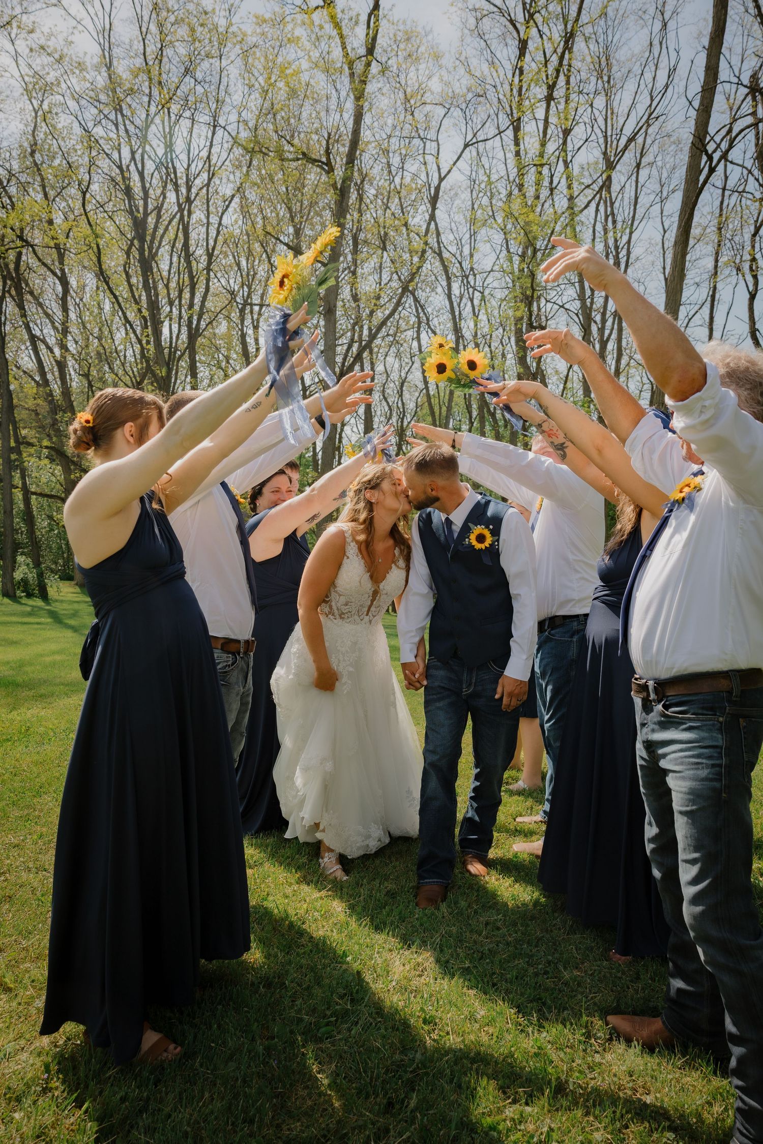 Bride and groom kiss under an arch of raised arms holding sunflowers, surrounded by wedding party in navy outdoors.