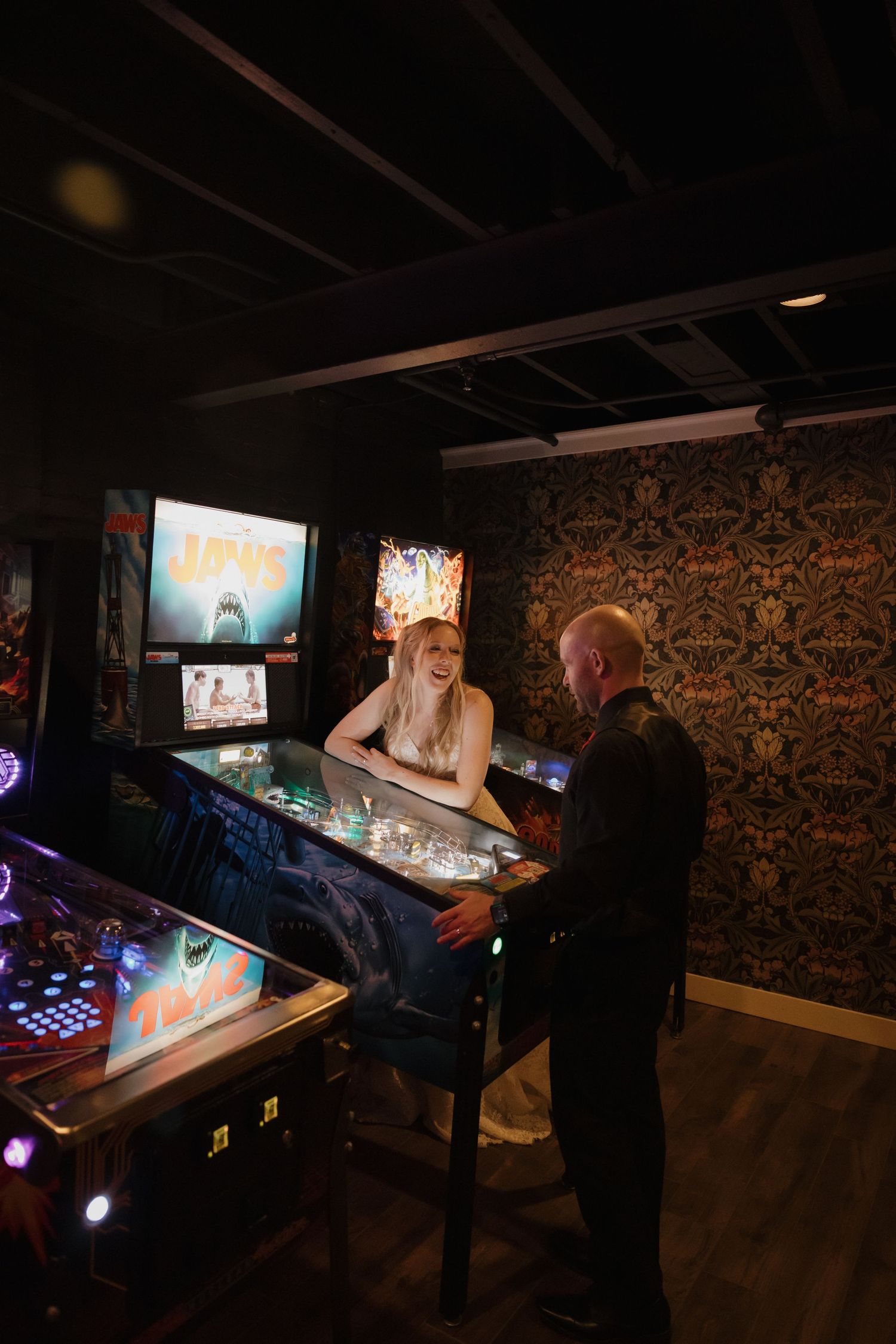 Couple laughing together at a Jaws pinball machine in a dimly lit vintage arcade game room.