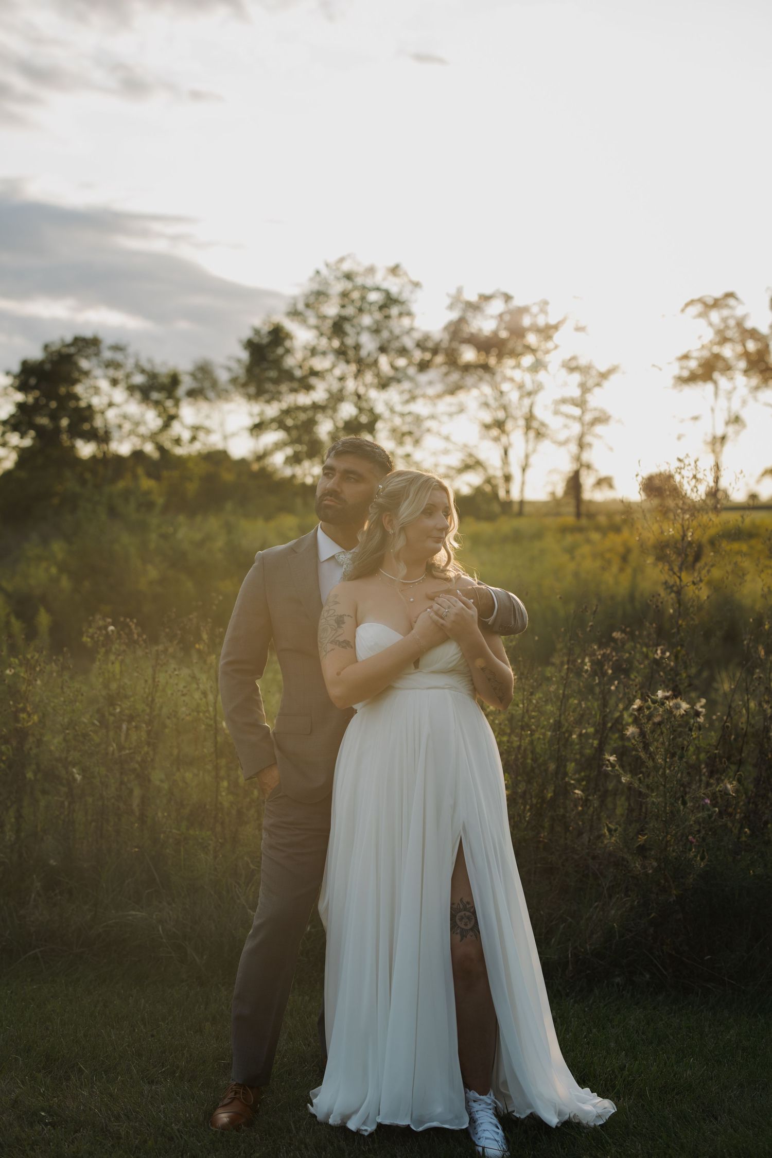 Newlyweds embrace in golden sunset light amidst natural field setting.