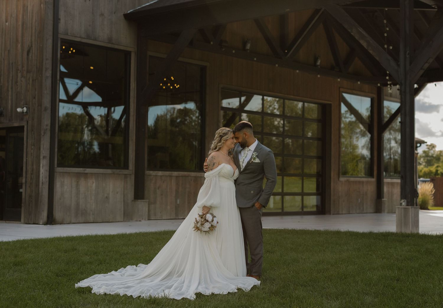 A couple in elegant wedding attire embraces under a rustic wooden barn structure during their special day.