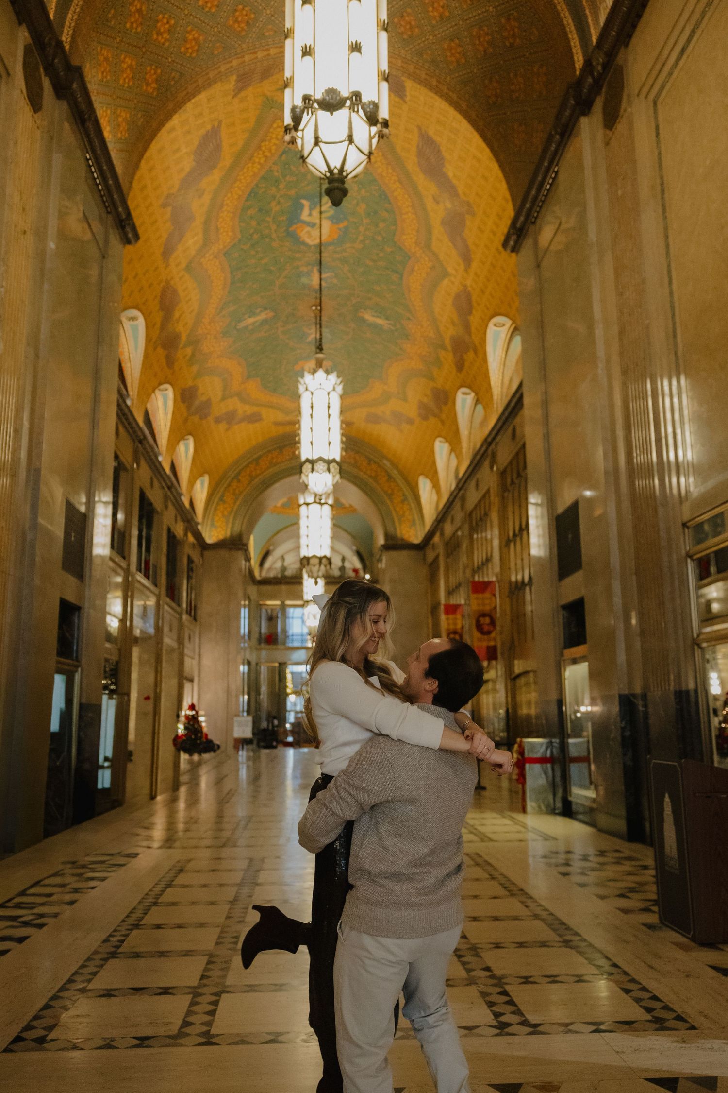 Couple embracing in ornate Art Deco hallway with golden mosaic ceiling and elegant chandeliers.