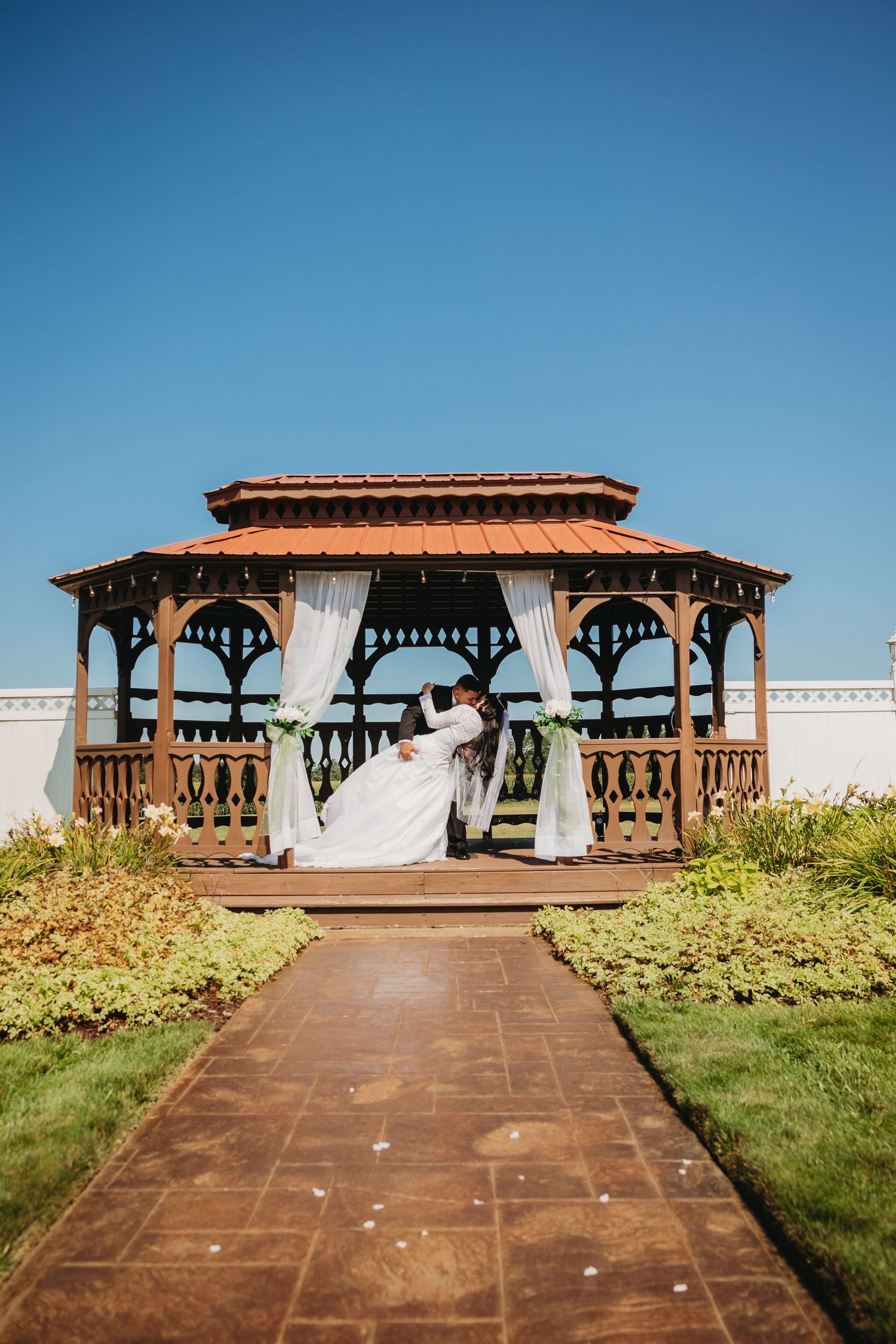Wedding ceremony setup at wooden gazebo with white draping and brick pathway.