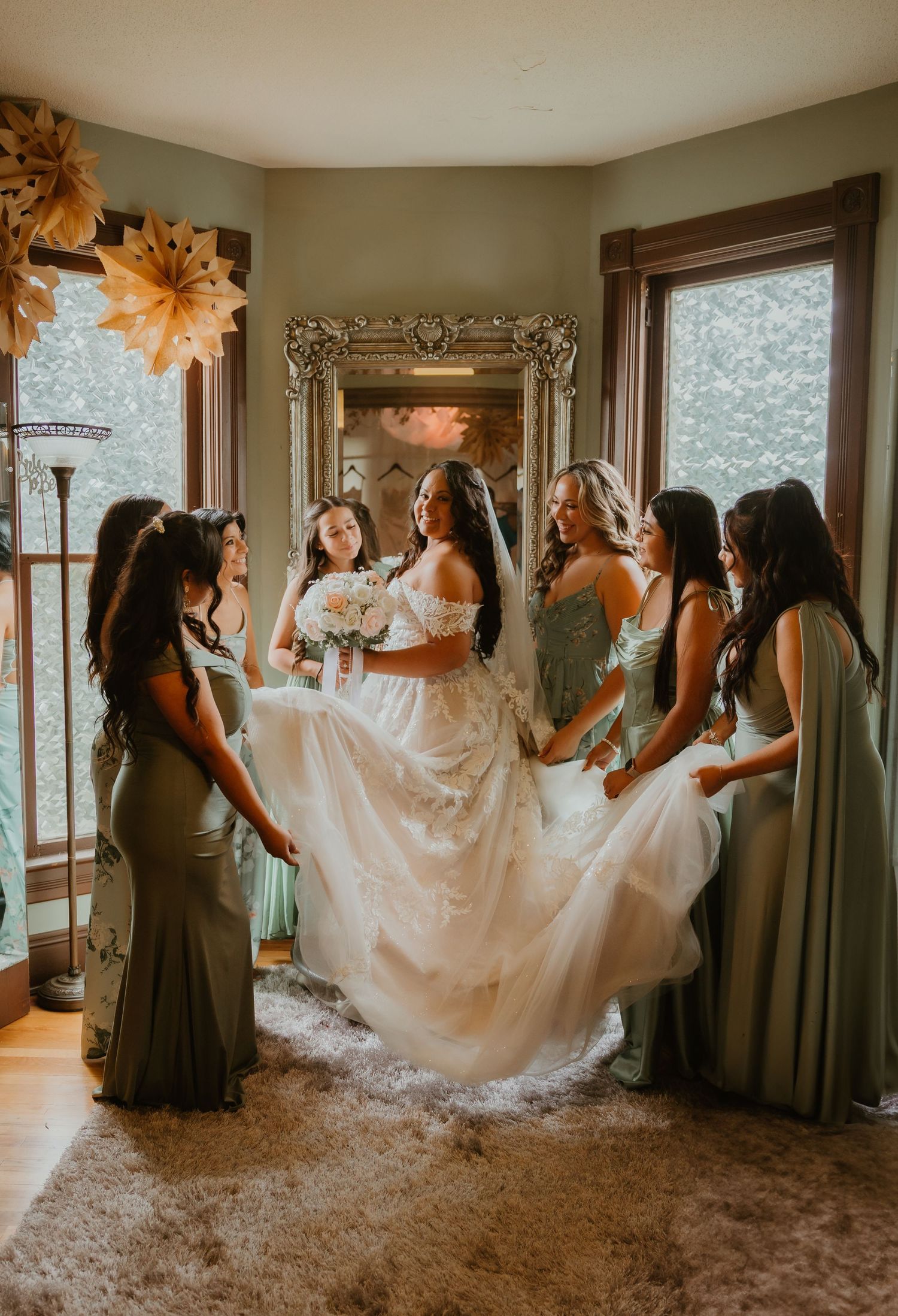 Group in elegant gray dresses assists with flowing white wedding gown preparations in a sunlit room.