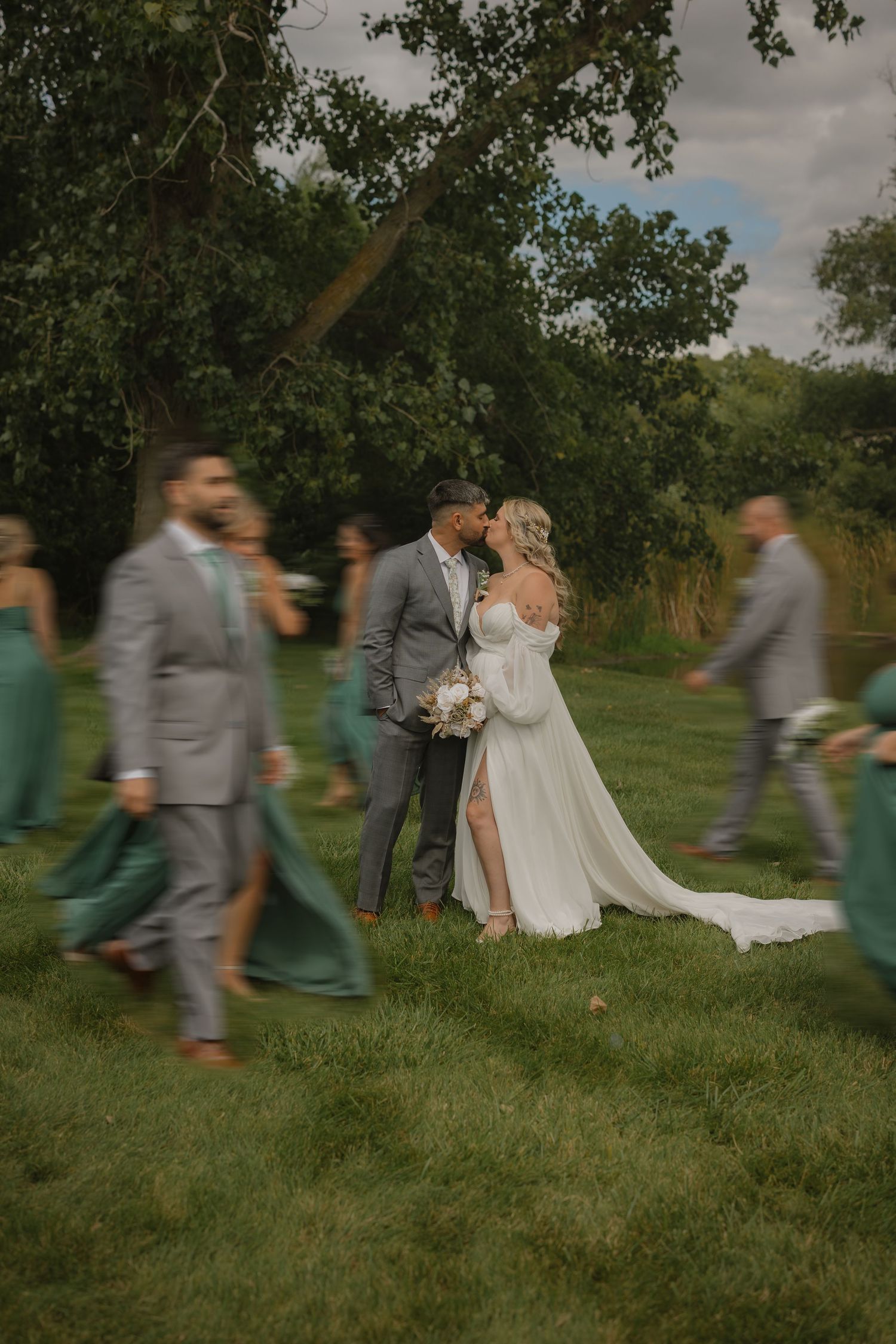 Wedding party members walk and dance across a grassy field in coordinated green and gray attire.