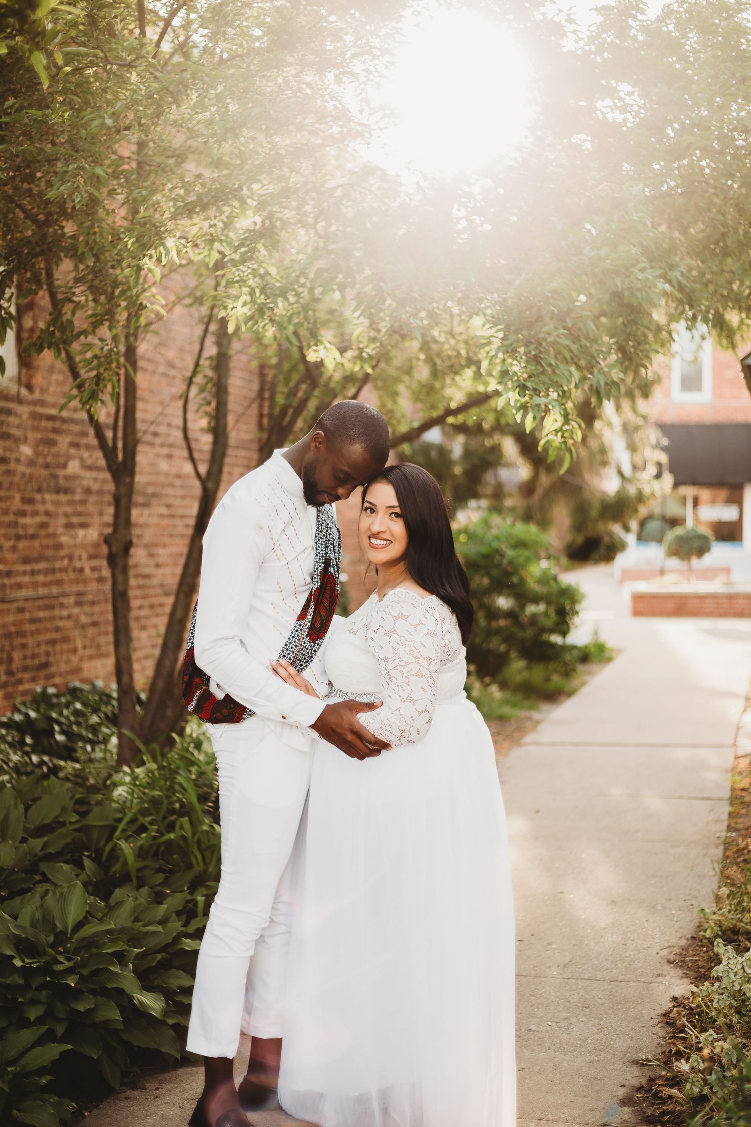 A couple in matching white outfits share joyful moments together on a sunlit path lined with greenery and brick walls.