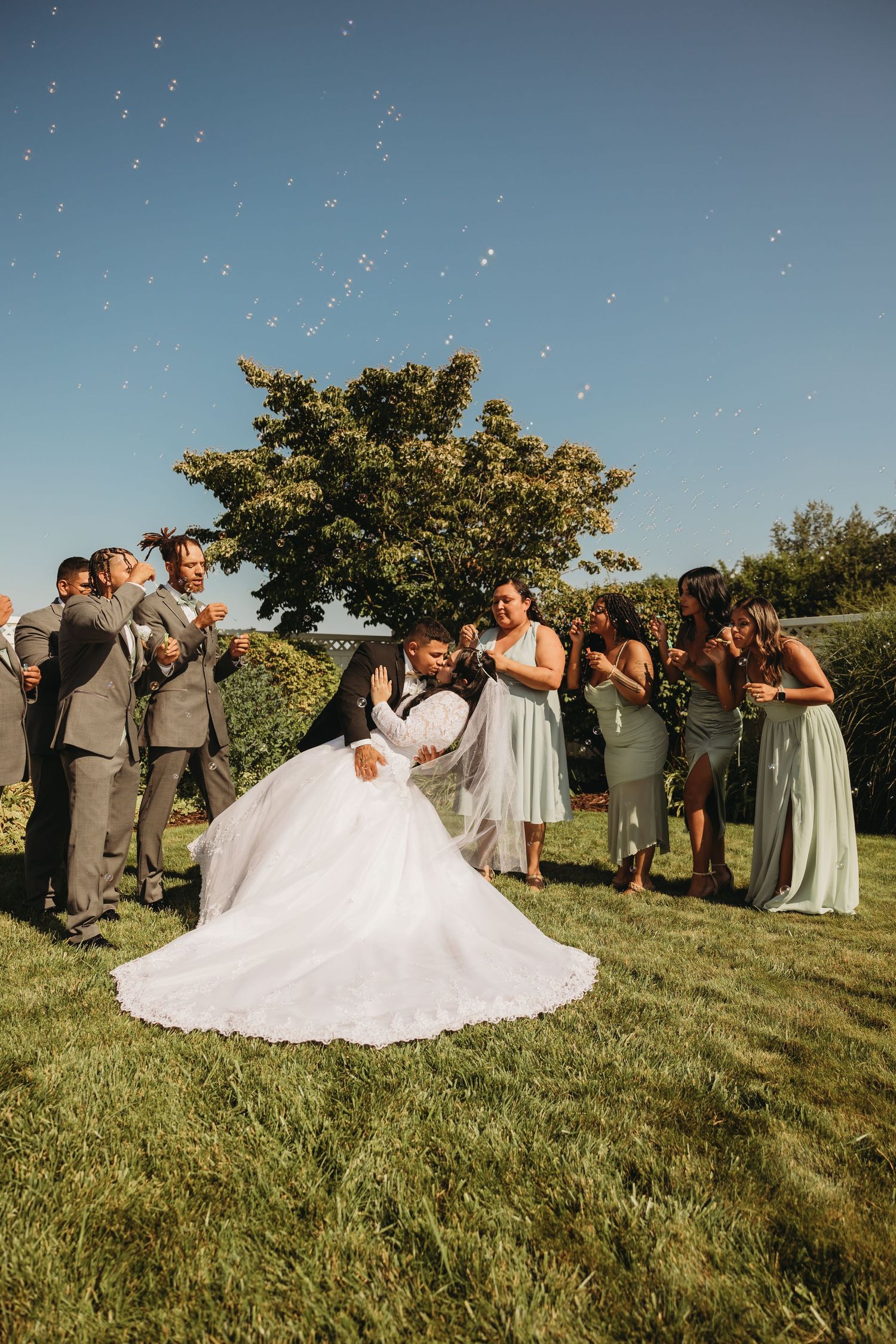 Wedding party celebrates on lawn with confetti and bubbles floating in blue sky.
