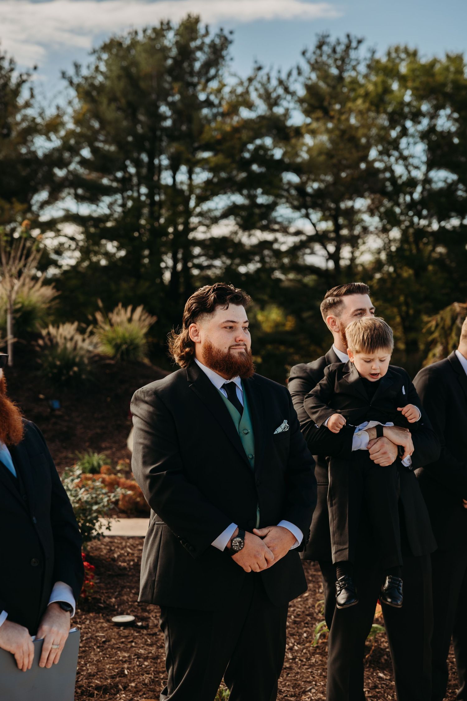 Groomsmen in formal suits stand outdoors during a wedding ceremony.