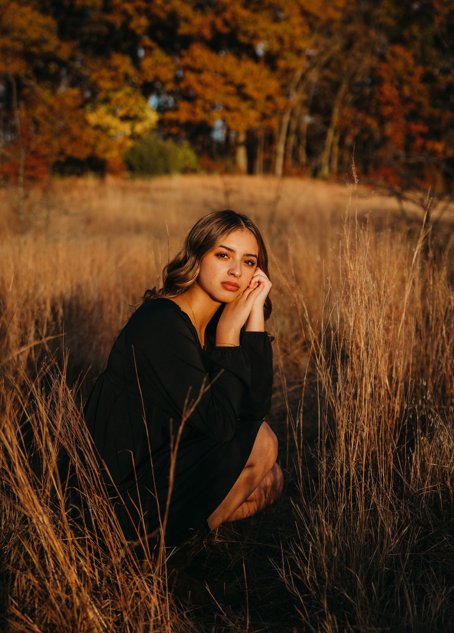 Young woman in black dress sitting in golden autumn field with colorful fall foliage trees in background.