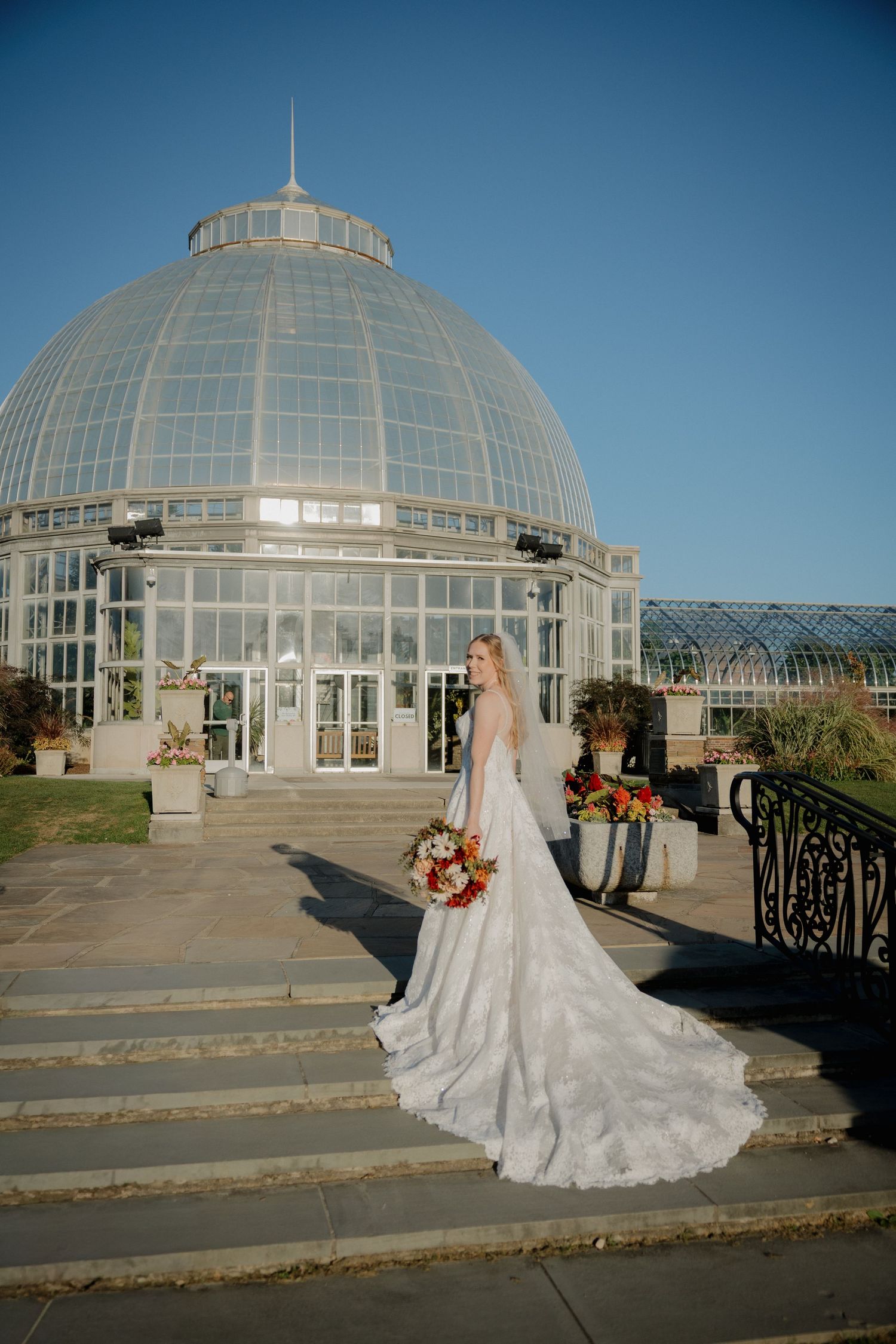Bride in lace gown with bouquet poses on steps before a grand glass dome conservatory.