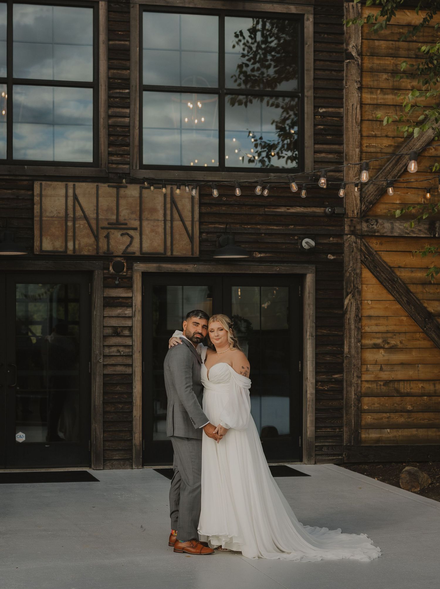 A wedding couple poses together outside a rustic wooden barn venue with string lights.
