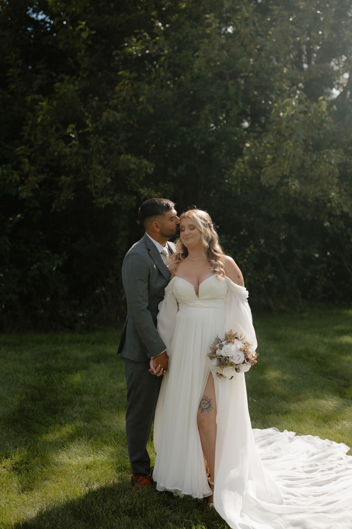 Romantic wedding couple embraces outdoors in a sunlit garden setting with flowing white dress and gray suit.