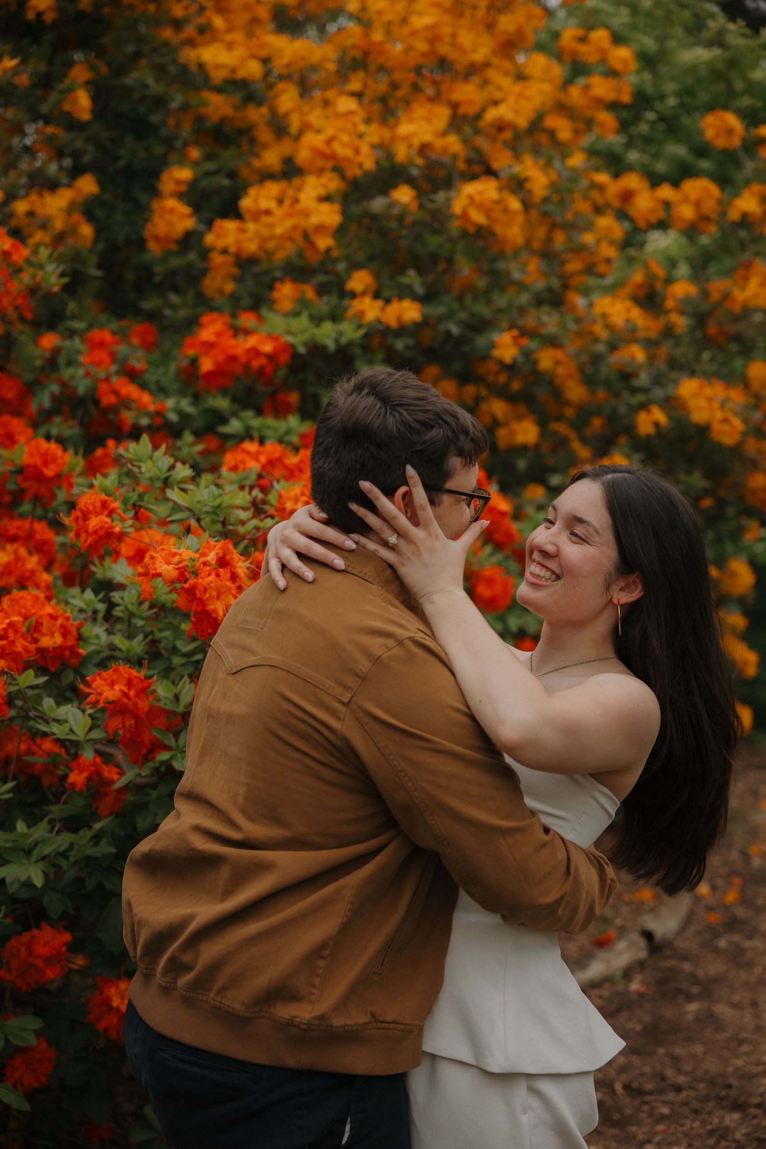 Couple embracing and smiling among vibrant orange and yellow flowering bushes during an outdoor engagement session.