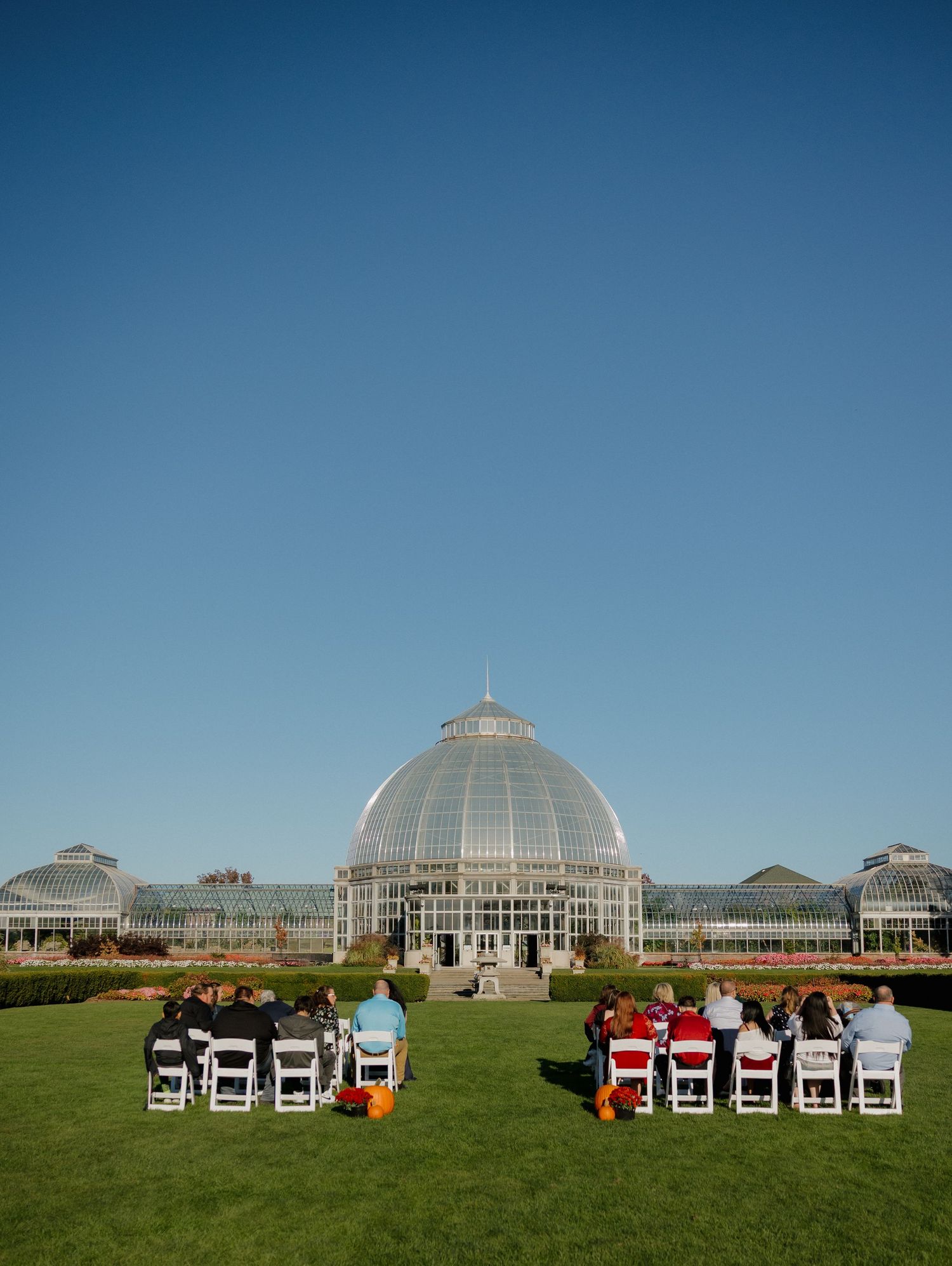 Small wedding ceremony on green lawn before grand glass-domed conservatory building under clear blue sky.