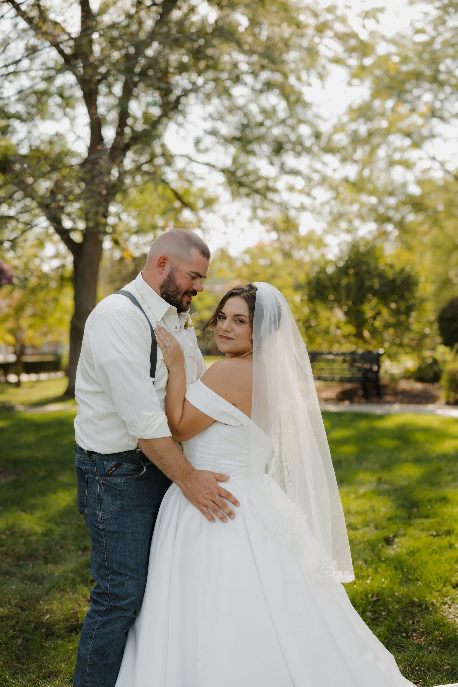 Bride in white gown and veil embraces groom in white shirt and jeans outdoors in a lush green park setting.