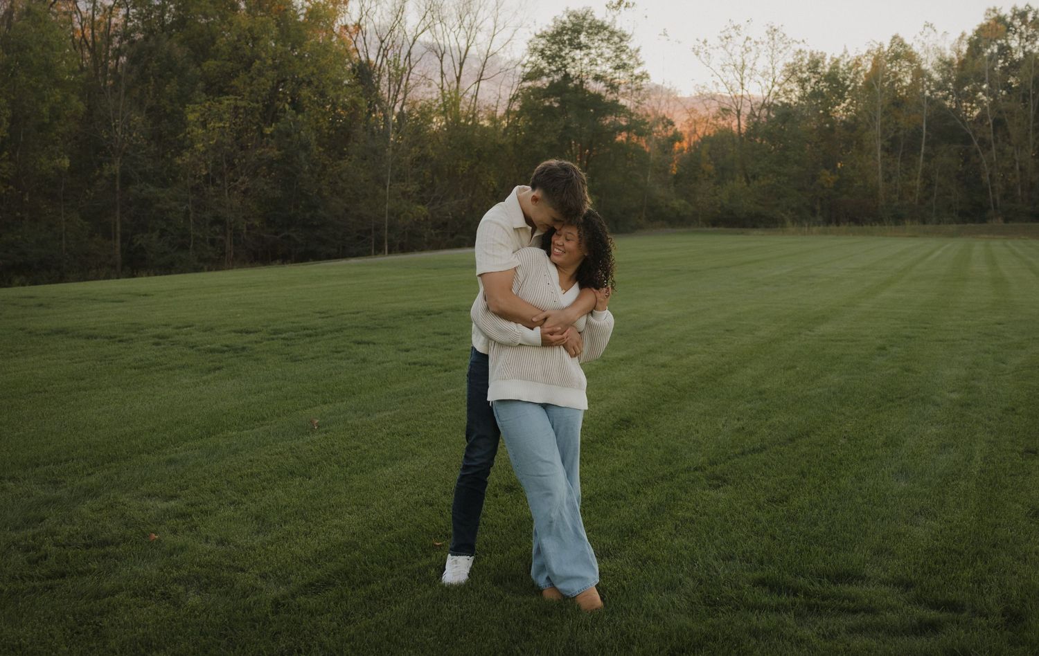 A couple embraces in a peaceful meadow during sunset with trees in the background.