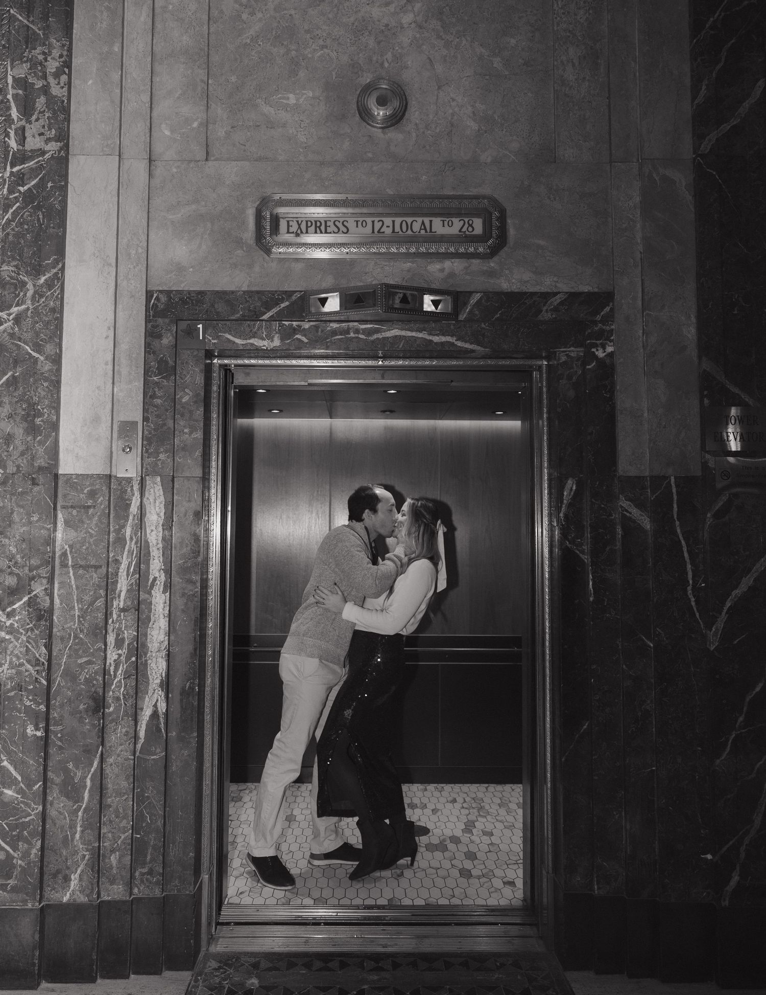 Black and white photo of a couple sharing a romantic kiss inside an elegant elevator with marble walls.