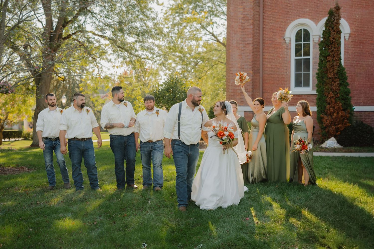 Wedding party poses outside brick church, groomsmen in jeans and suspenders, bridesmaids in sage green dresses, bride holds floral bouquet.
