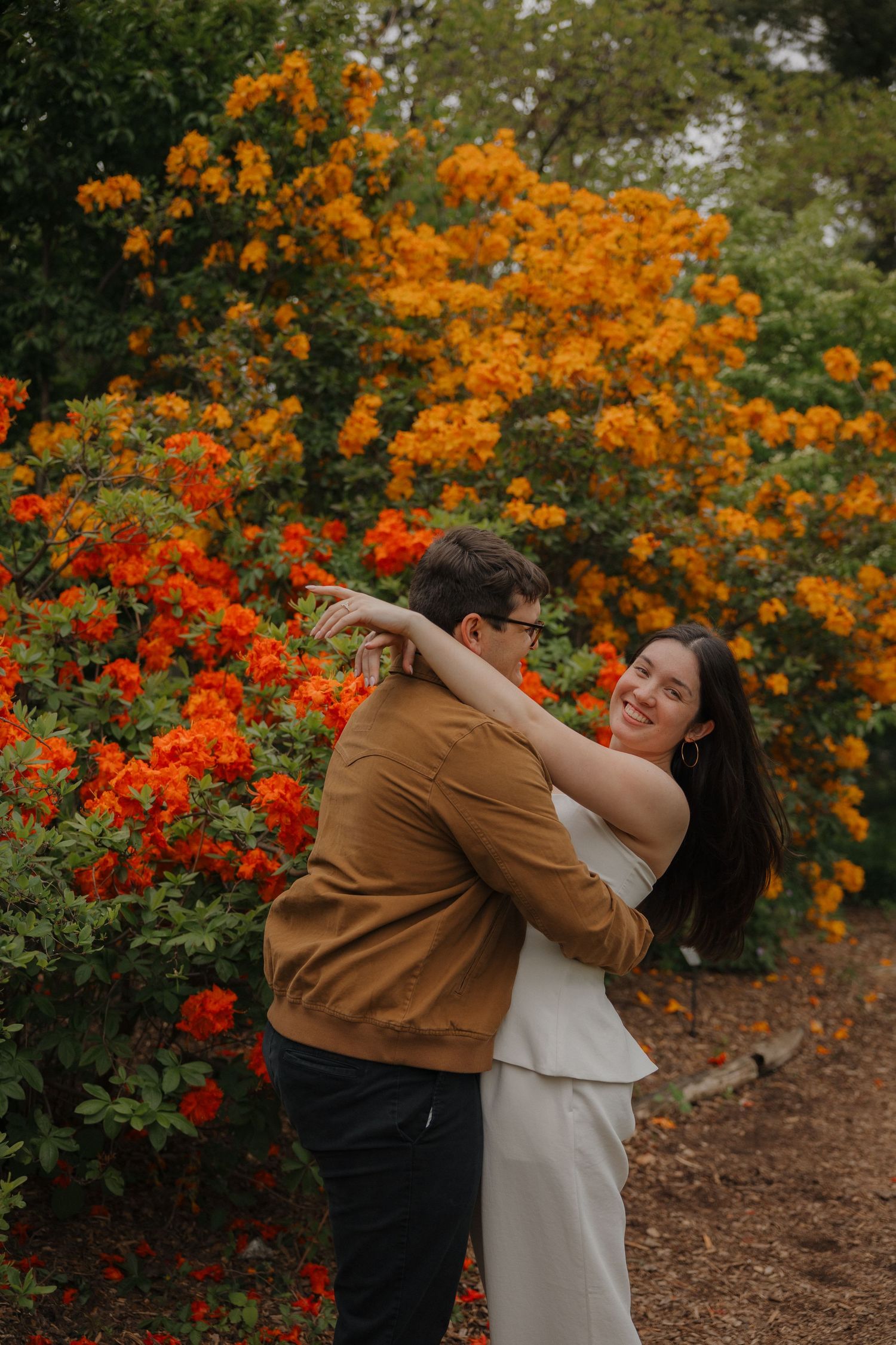 Couple embracing and smiling in front of vibrant orange and red flowering bushes in a garden.
