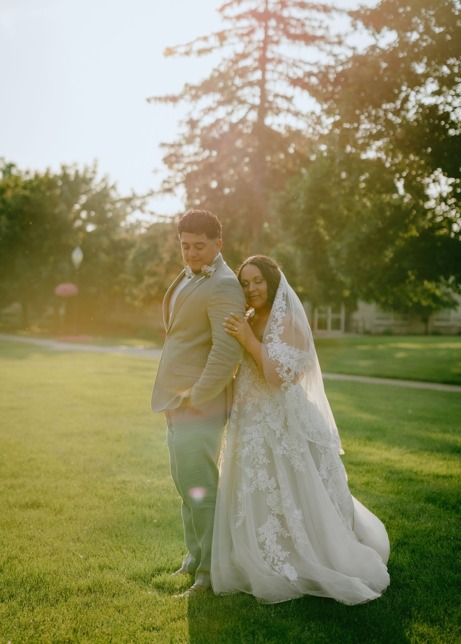 A newlywed couple walks together on a sunlit green lawn during golden hour at their wedding celebration.