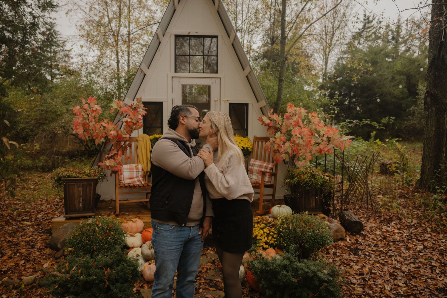 Romantic autumn engagement photoshoot with couple embracing among fallen leaves and rustic white house backdrop.