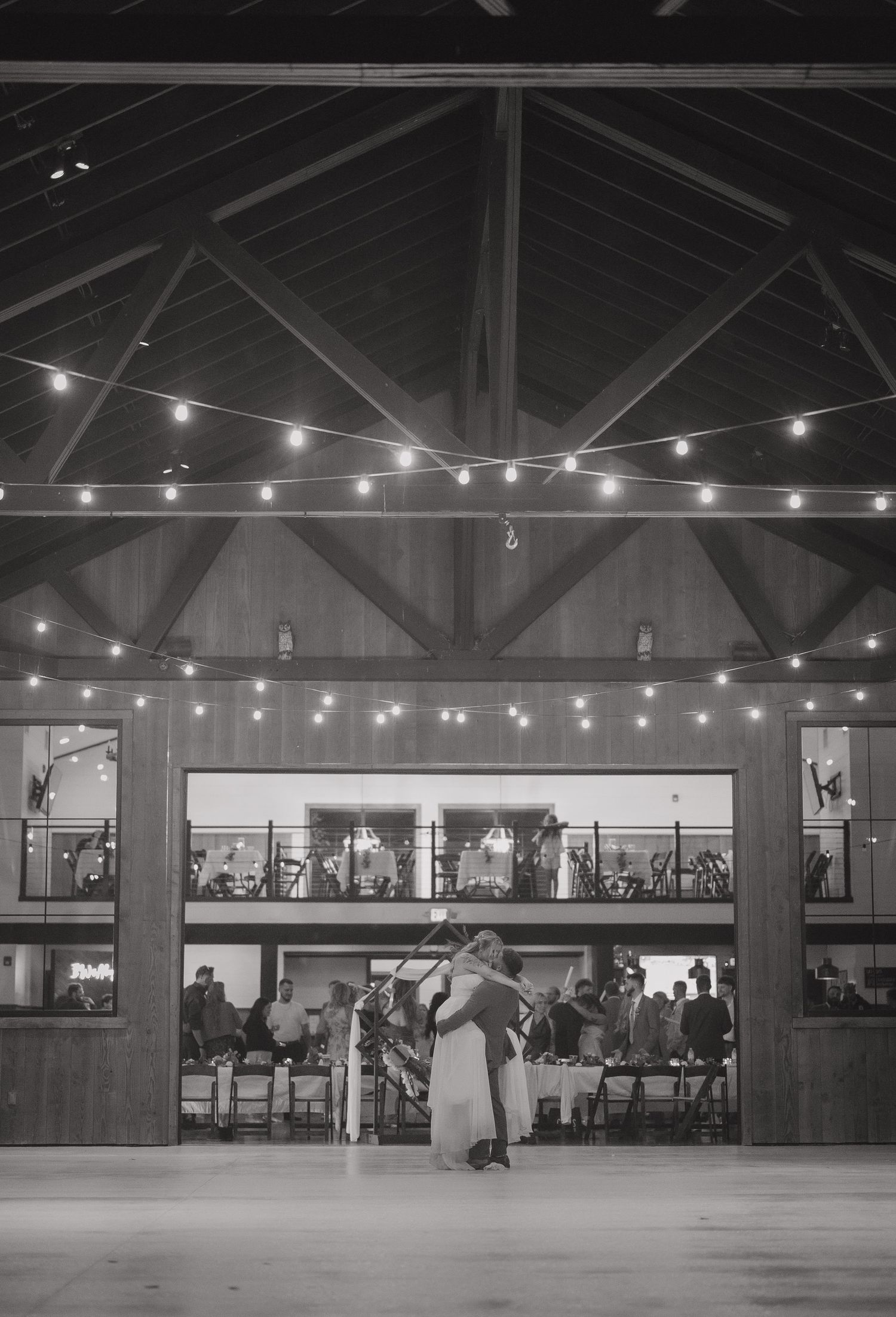 A black and white photo of a wedding venue with string lights and exposed wooden beams.