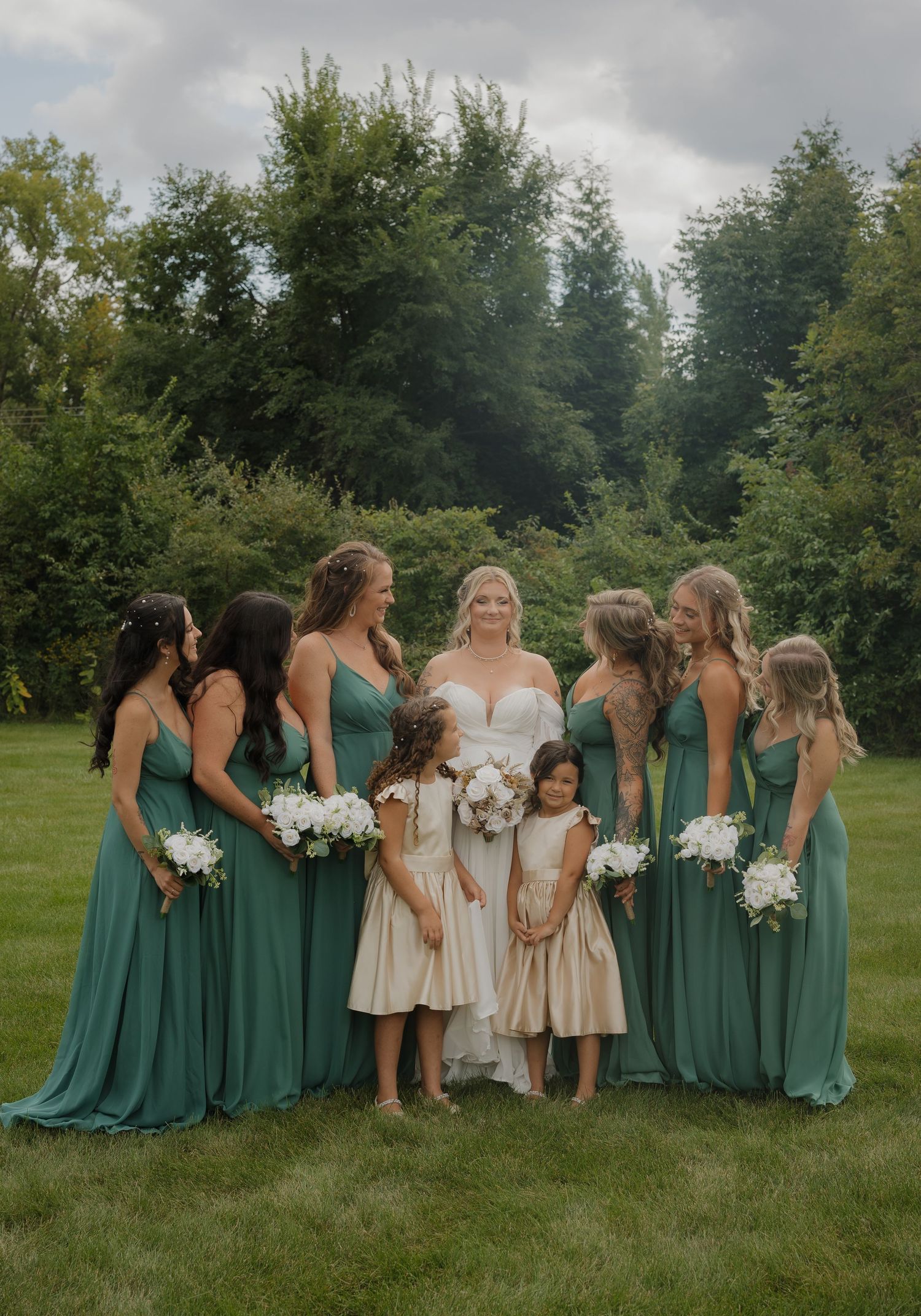 Bridesmaids in emerald green dresses and flower girls in cream dresses pose on grass for wedding photos.
