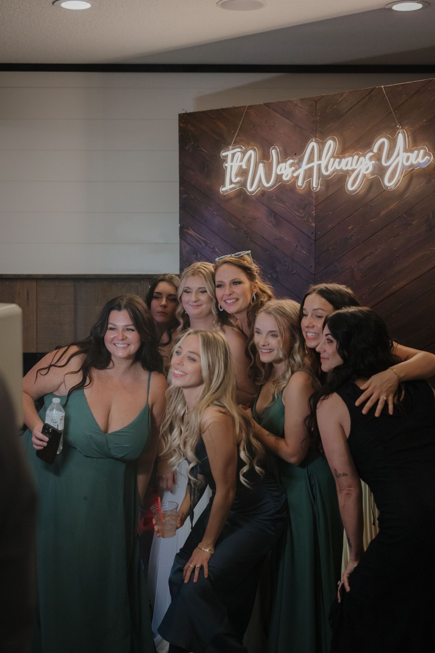 A group of bridesmaids pose together under a neon sign reading 'I'll Always Love You'.