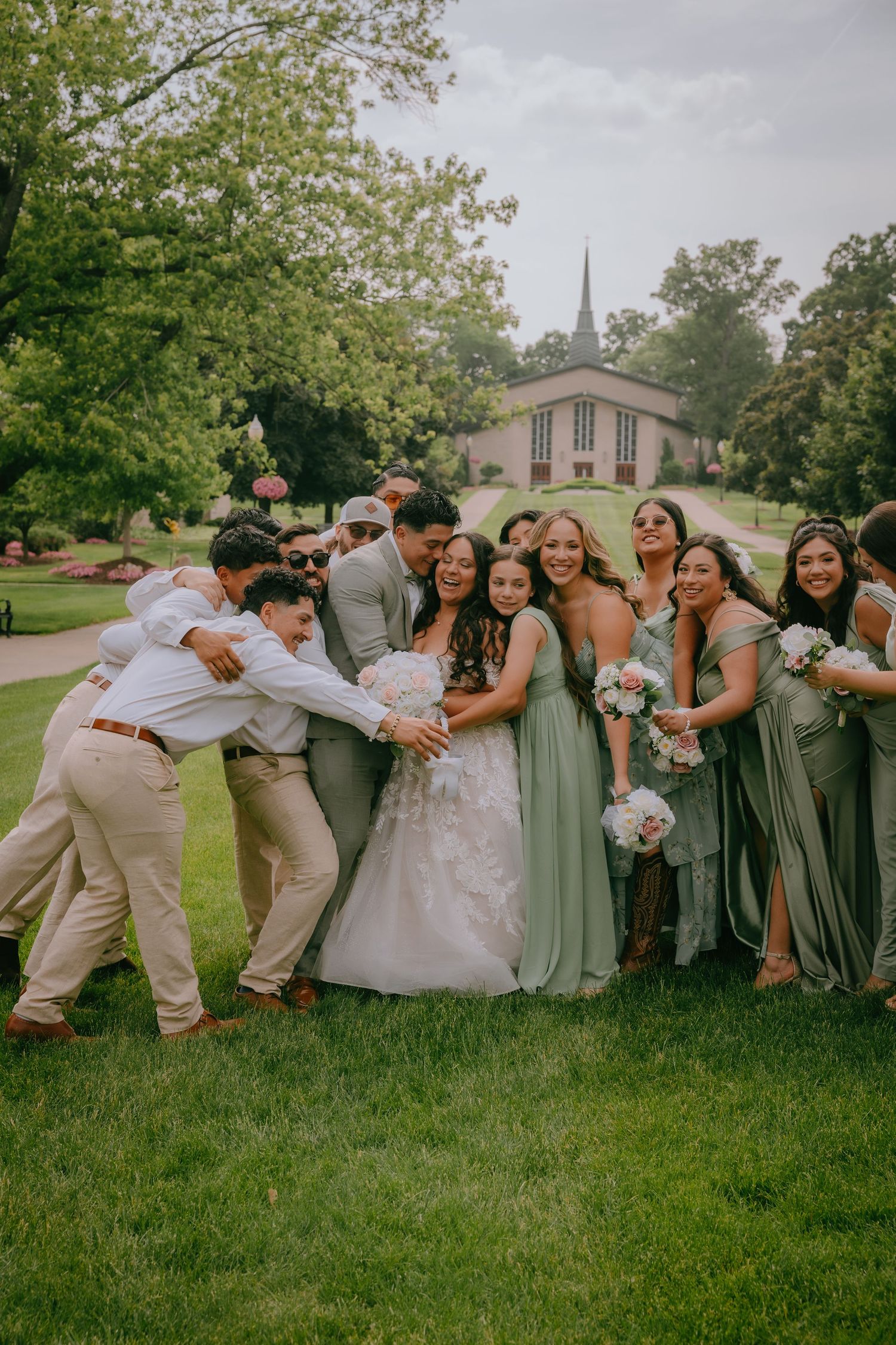 Wedding party in sage green dresses and khaki suits playfully posing together on a lawn.