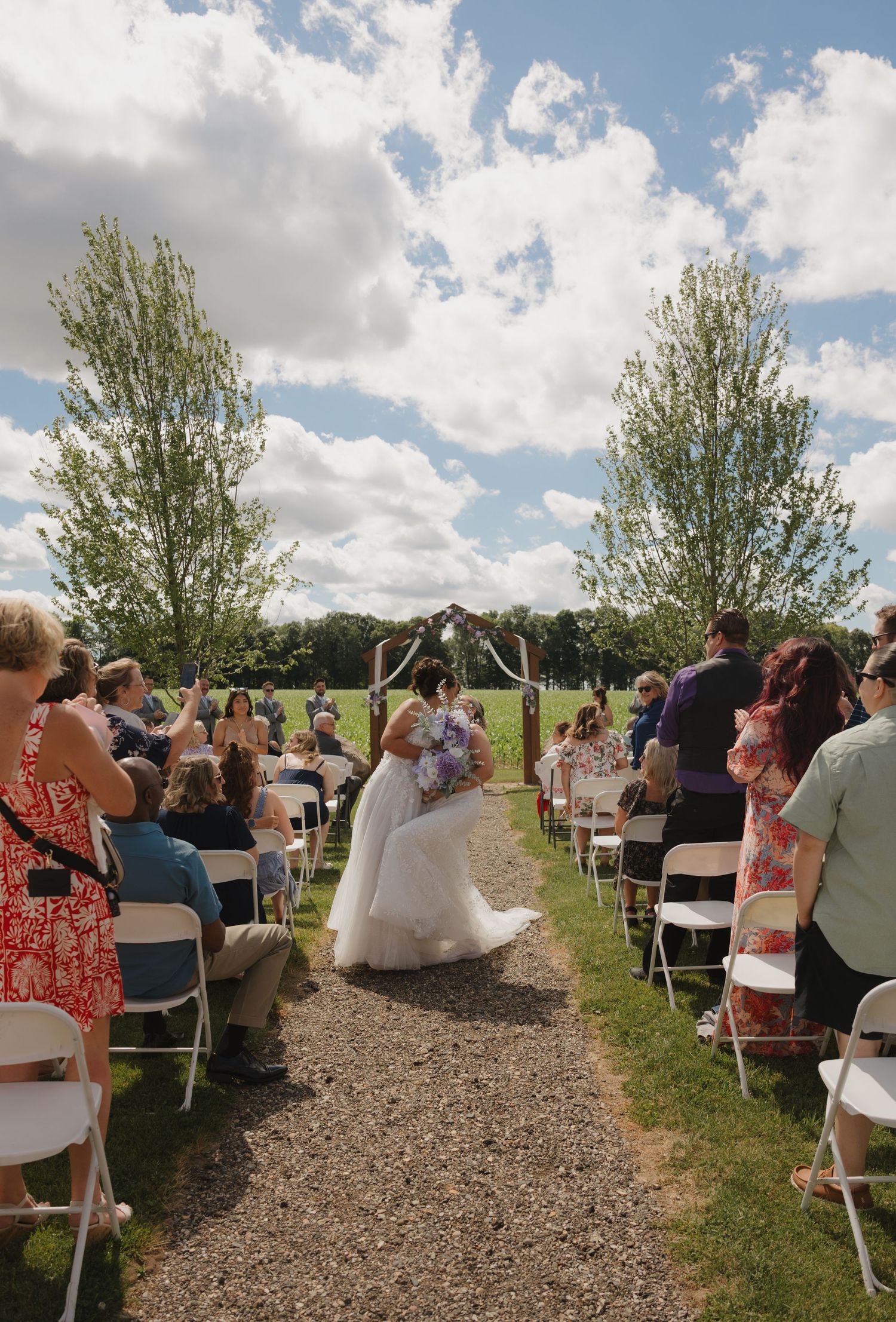 Bride walks down outdoor wedding aisle lined with white chairs and trees under a blue sky.