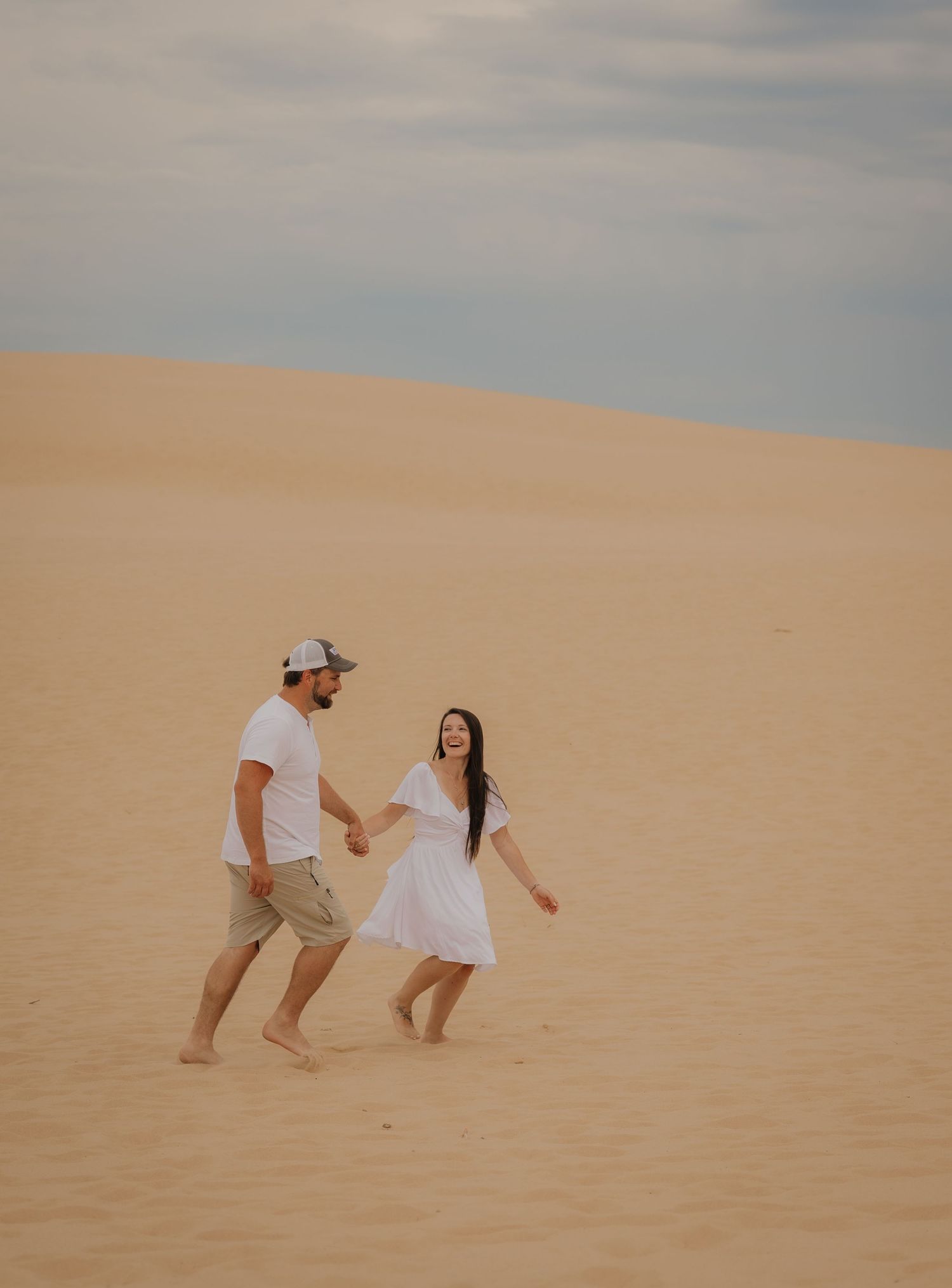 A couple holds hands and walks barefoot across golden sand dunes under a cloudy sky, woman in white dress.