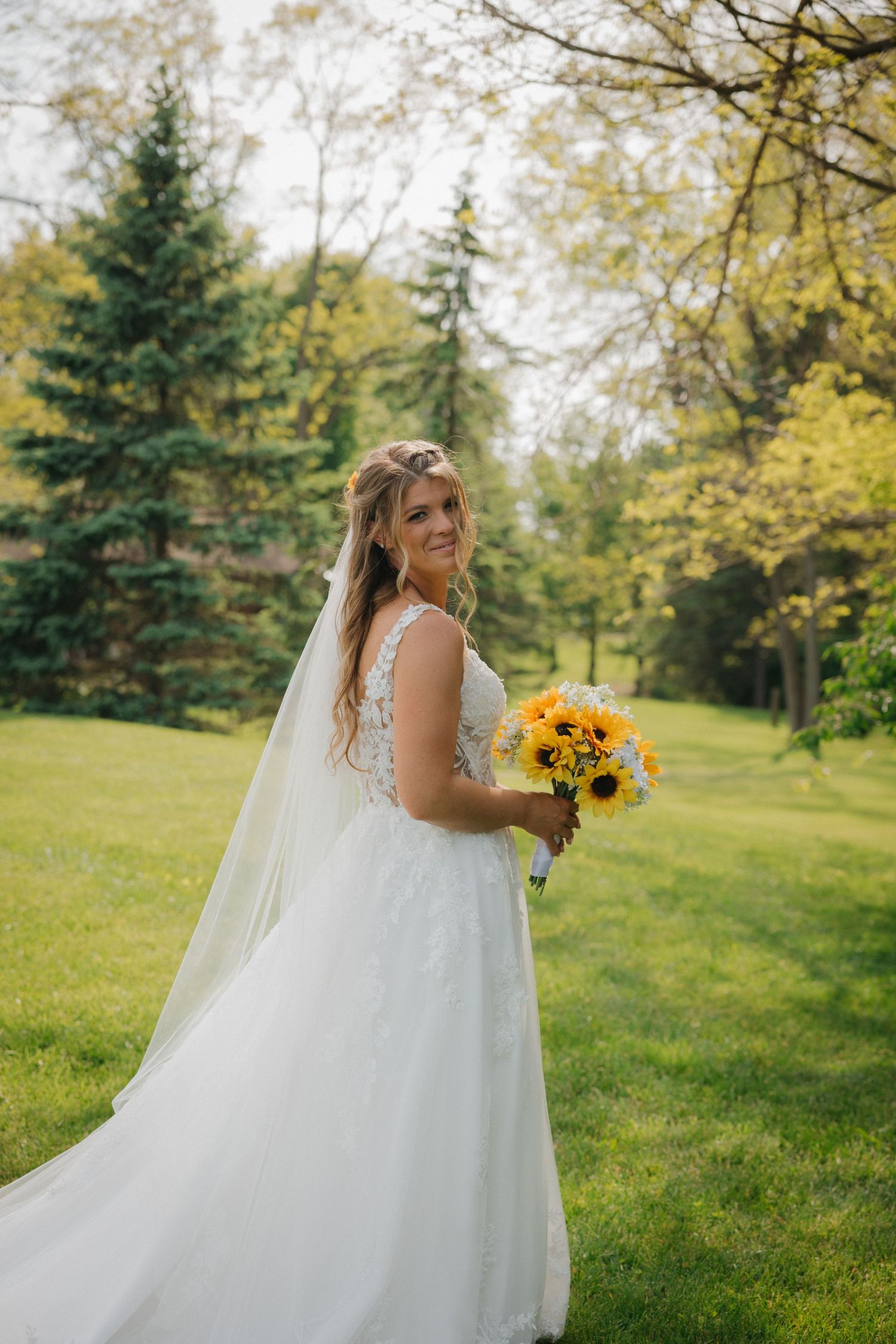 Bride in white lace gown with veil holds sunflower bouquet outdoors on green lawn surrounded by trees.