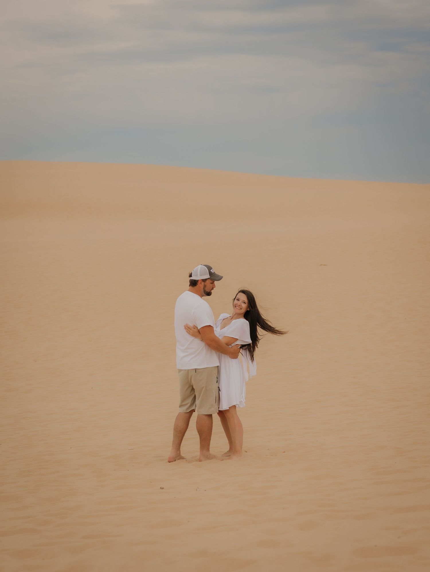 Couple embracing on sandy desert dunes, woman in white dress with hair blowing, man in white shirt and cap.