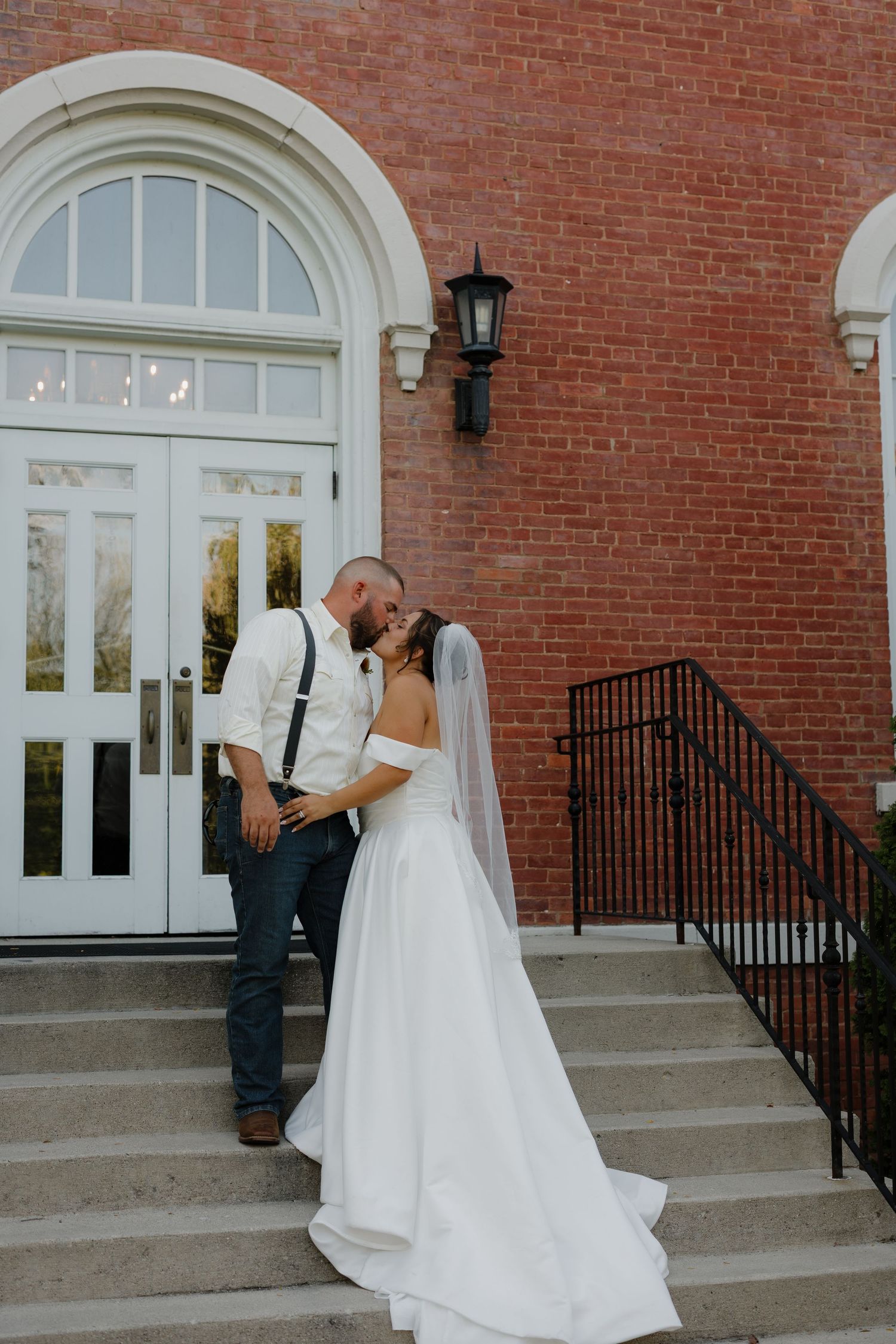 Bride and groom share a romantic kiss on brick building steps, bride in white gown with veil, groom in jeans and suspenders.
