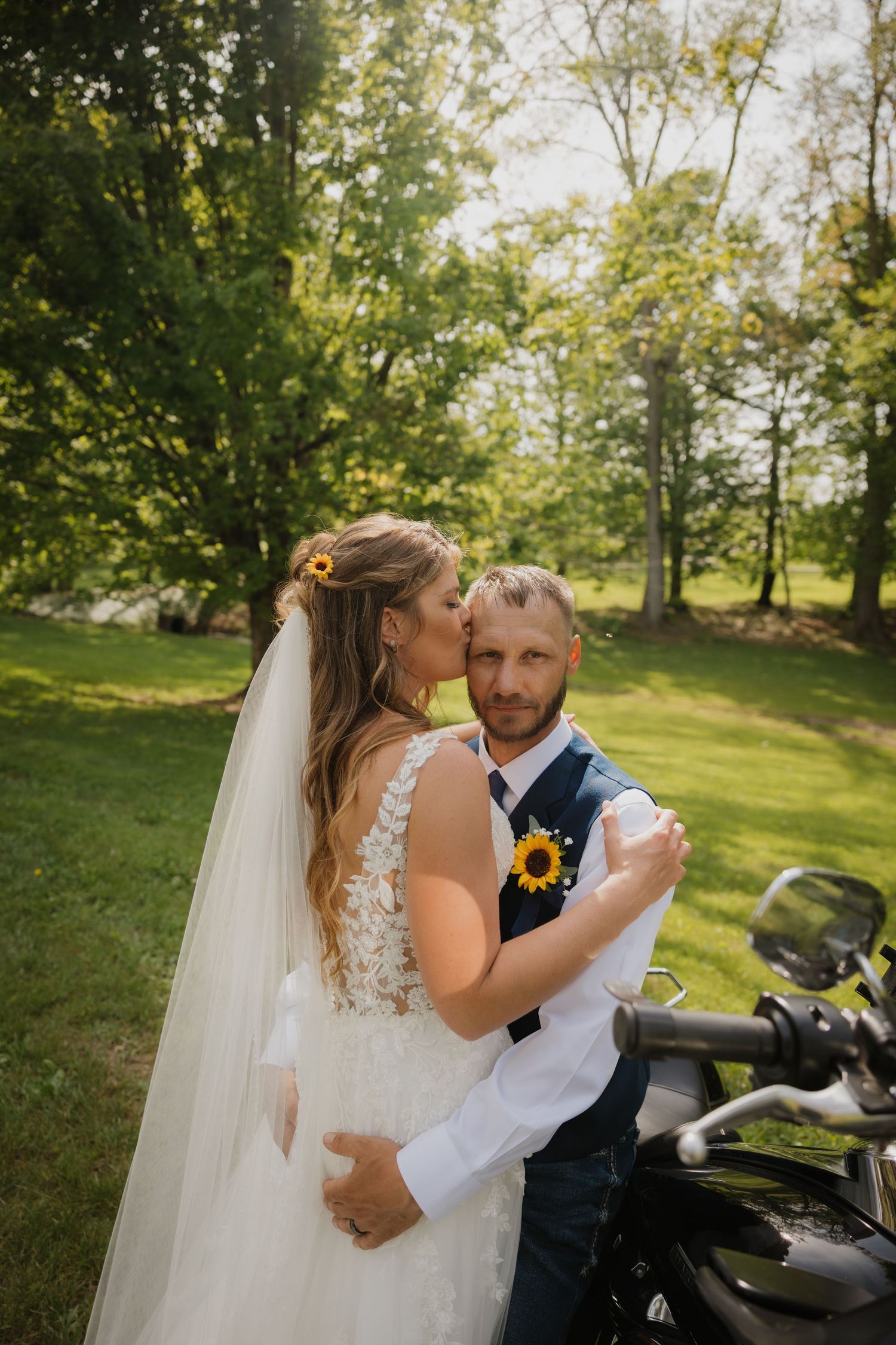 Bride kisses groom's cheek as they dance outdoors near a motorcycle, both wearing sunflowers, in a lush green park setting.