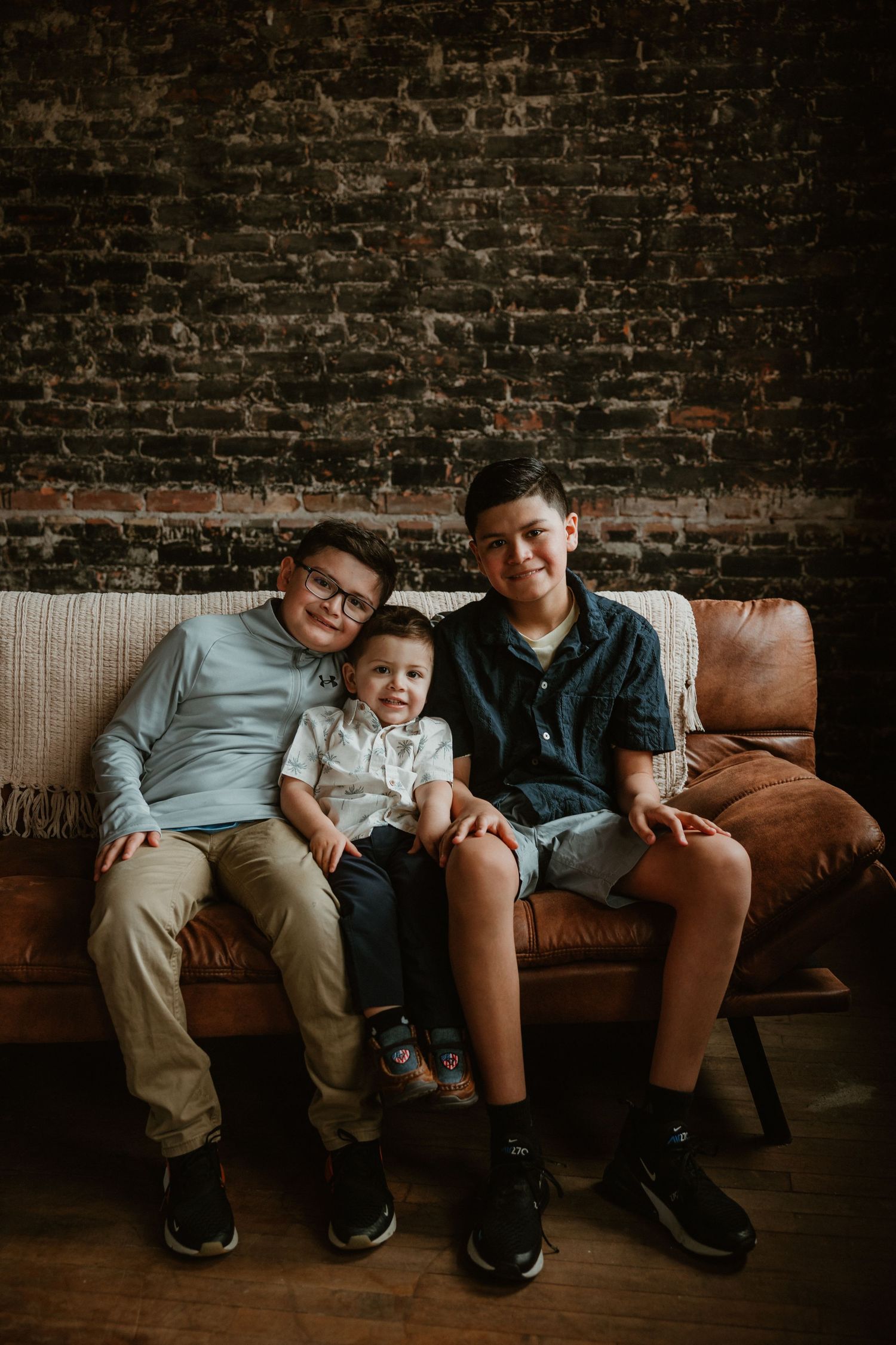 Three people seated on a leather couch against exposed brick wall in a cozy indoor setting.