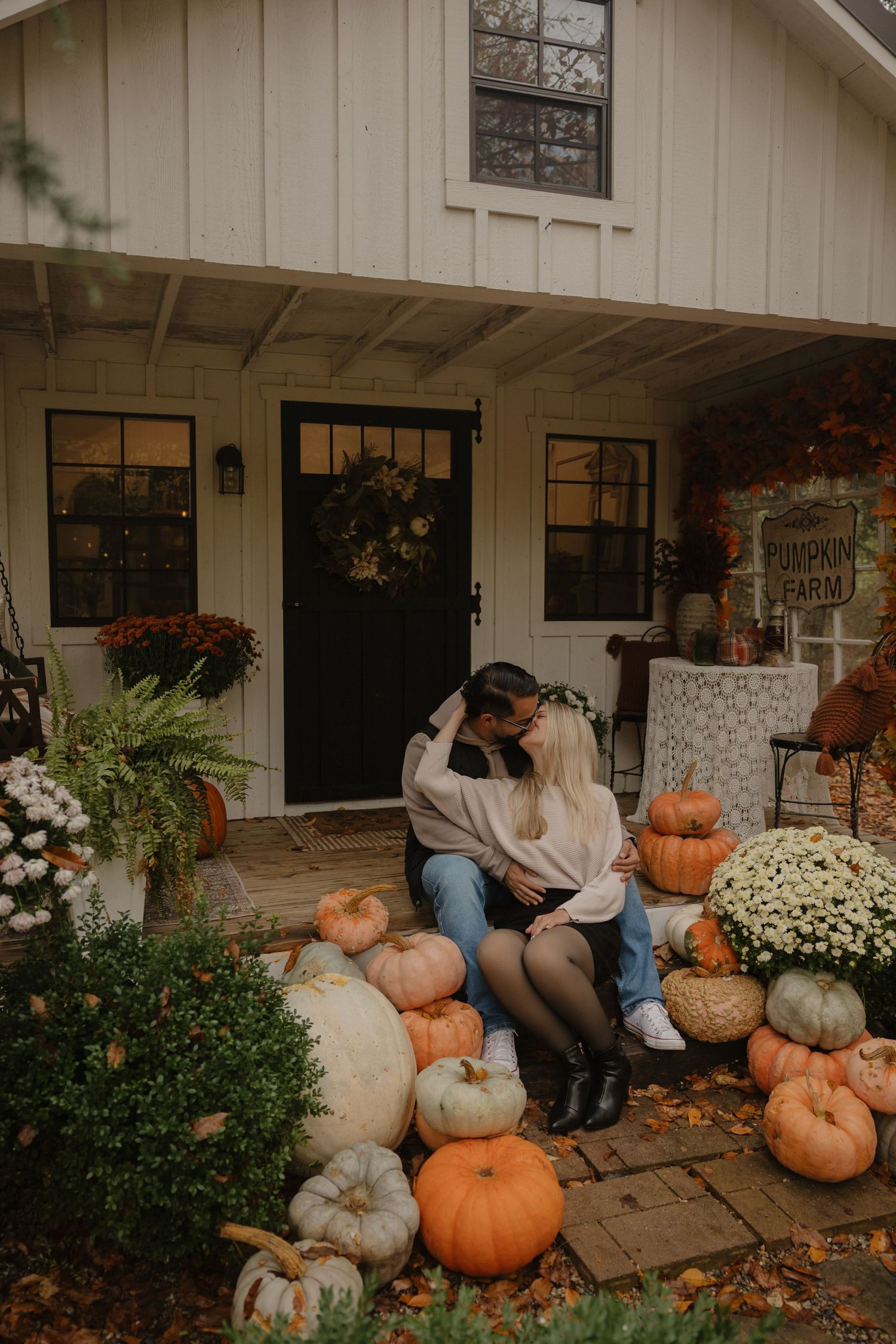 Autumn scene with pumpkins and mums decorating a white farmhouse porch with people sitting on stone steps.